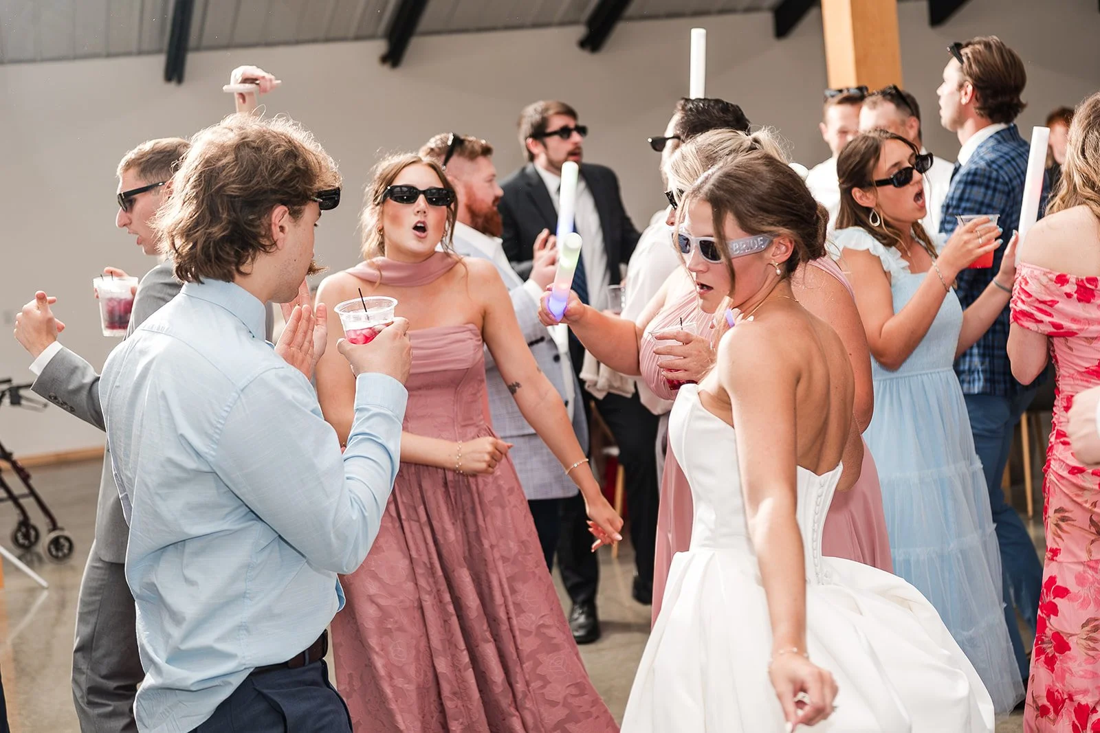 People dancing and celebrating at a wedding reception with the bride and groom in the center, surrounded by guests in colorful dresses and formal wear in a decorated ballroom.