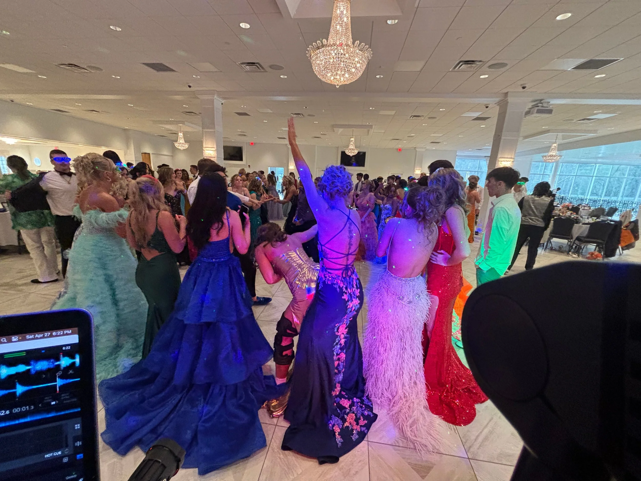 A dance floor filled with people in formal attire, including women in colorful dresses and men in suits, at an indoor event with chandeliers and banquet tables in the background.