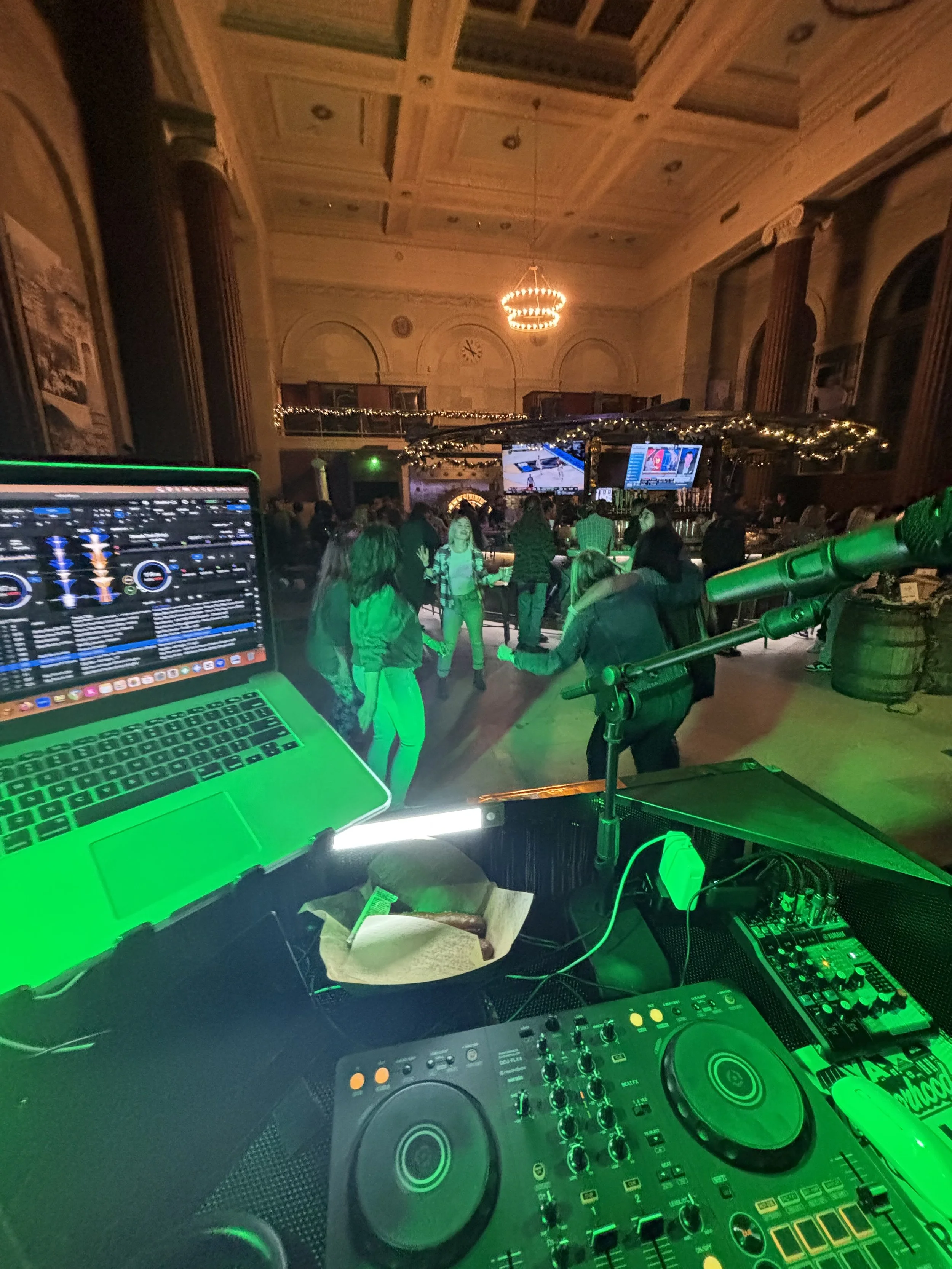 View from DJ's booth overlooking a dance floor with people dancing at a party or event in an elegant, decorated hall with chandeliers and large windows.