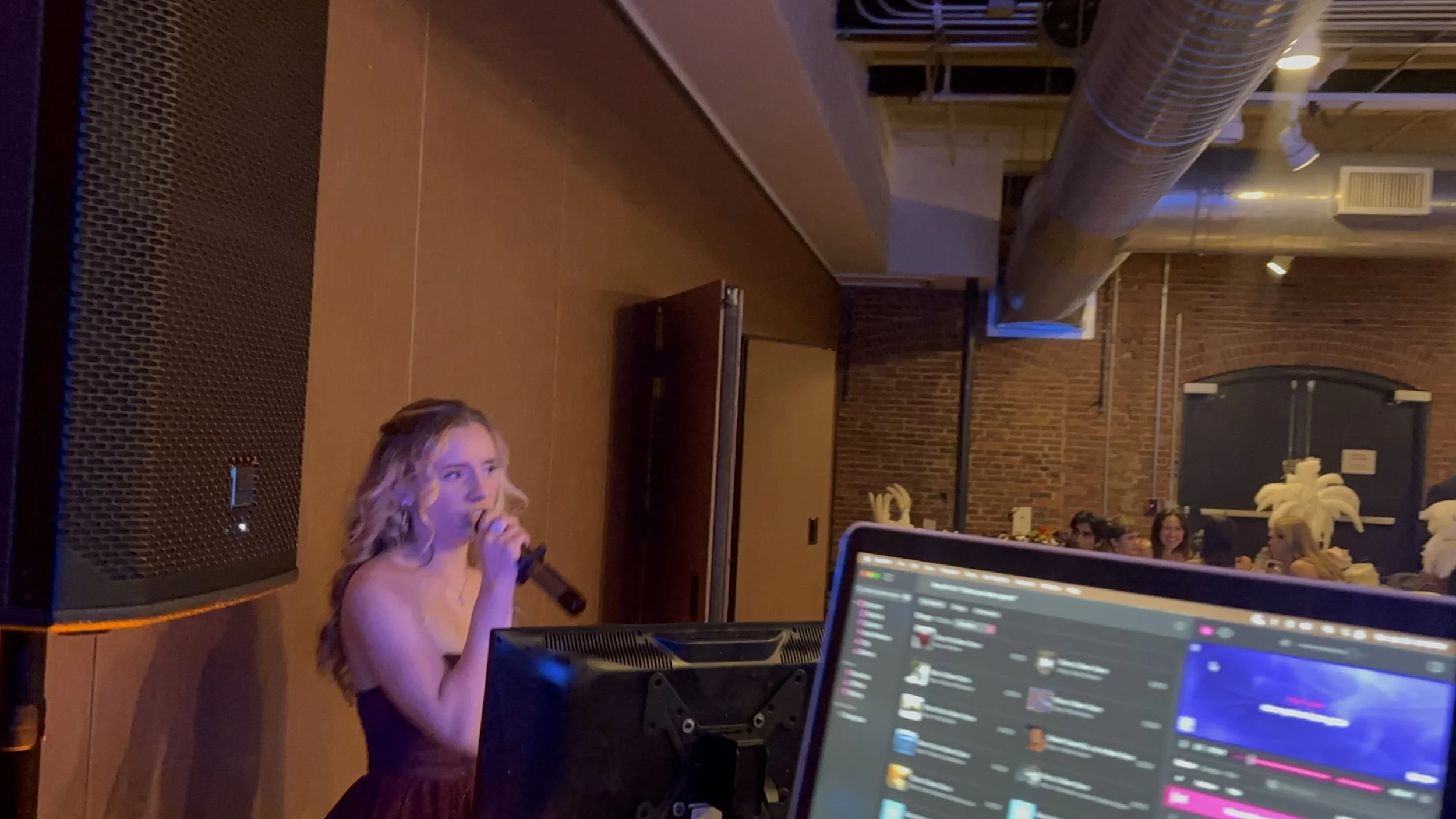 A woman with long curly blonde hair singing into a microphone at an indoor event, with a computer screen showing Discord open in the foreground and a group of people smiling in the background.
