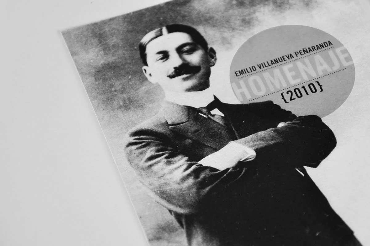 Black-and-white photo of Emilio Villanueva Peñaranda with glasses and mustache, crossing his arms, wearing a suit and tie. There is a pink circular label with his name, the text "HOMENAJE," and the date "02010".