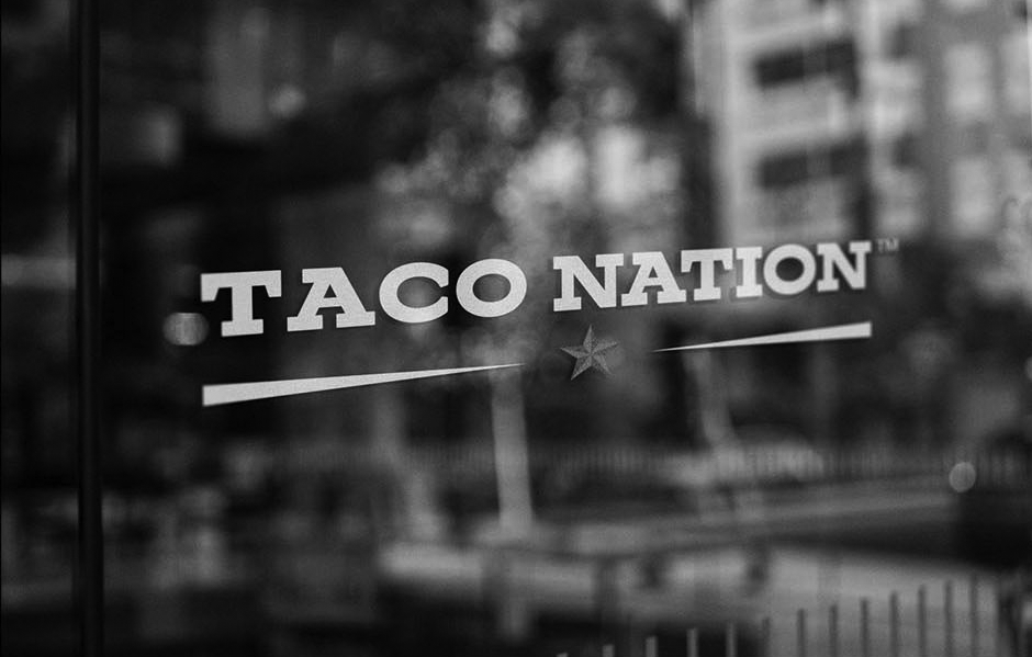 The glass door of a restaurant or food establishment displays the 'Taco Nation' logo with white text, a red star, and a white underline, with a blurred urban street scene reflecting in the glass.
