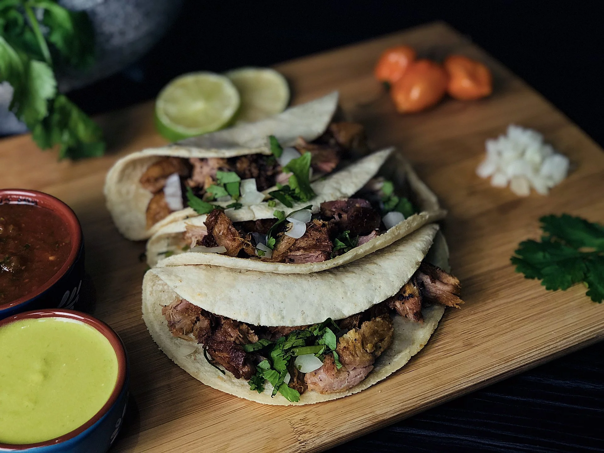 Two tacos filled with shredded meat, chopped onions, and cilantro, served on a wooden cutting board with lime wedges, chopped onions, diced tomatoes, and garnishes, alongside two small bowls of salsa and sauce.