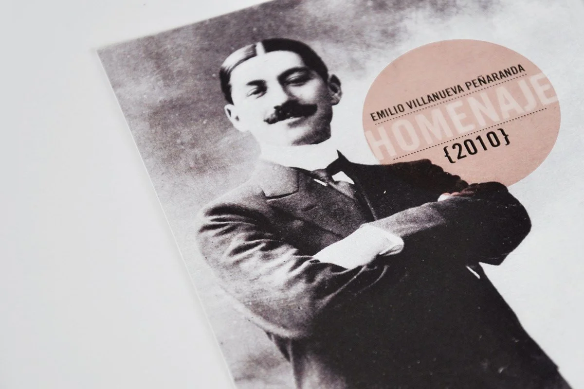 Black and white photograph of Emilio Villanueva Peñaranda with a mustache, wearing a suit with arms crossed. There is a pink circular label overlay containing his name, the title 'Home Naje,' and some numbers.
