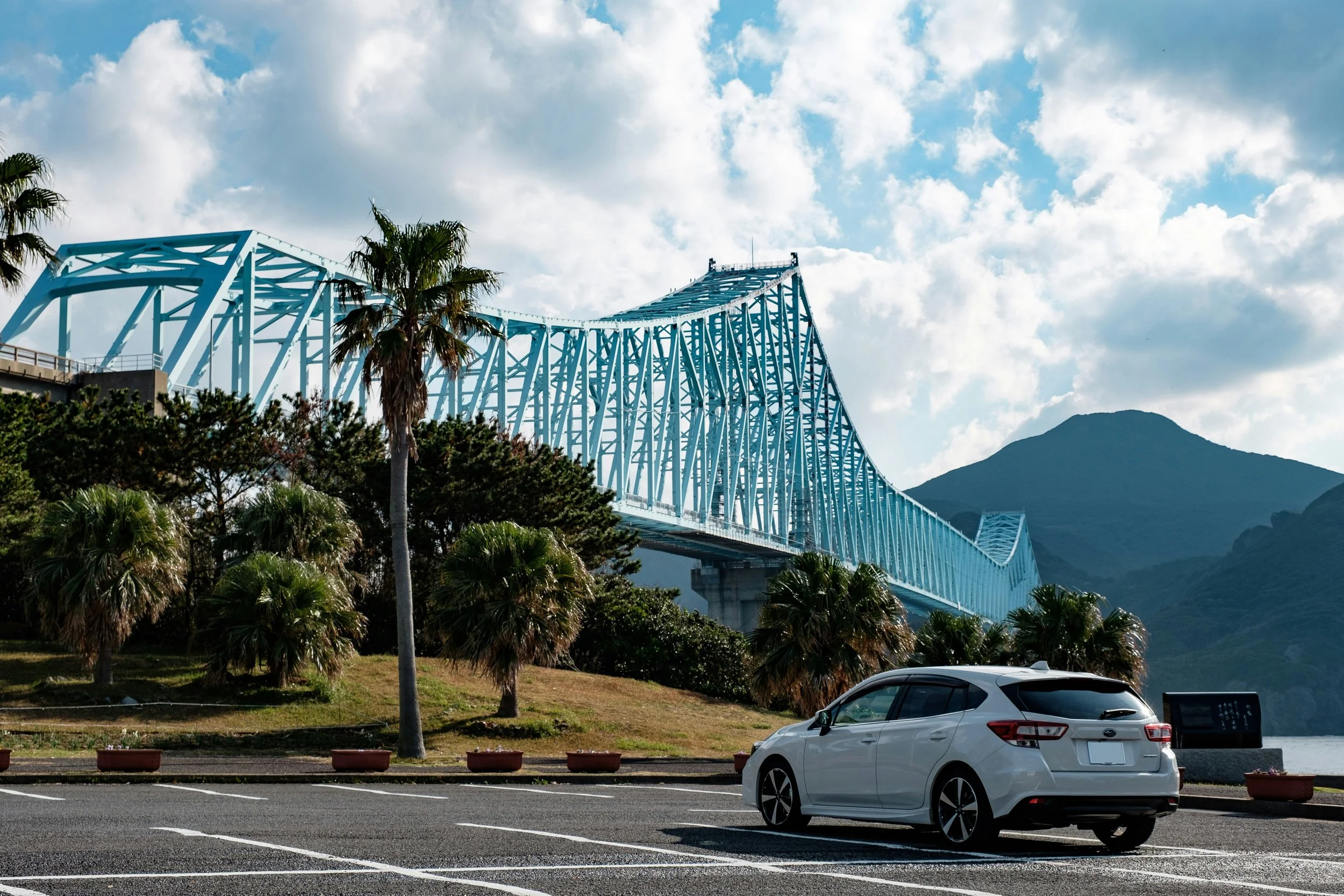 Hatchback car in Hirado, Japan parks under a palm tree and bridge