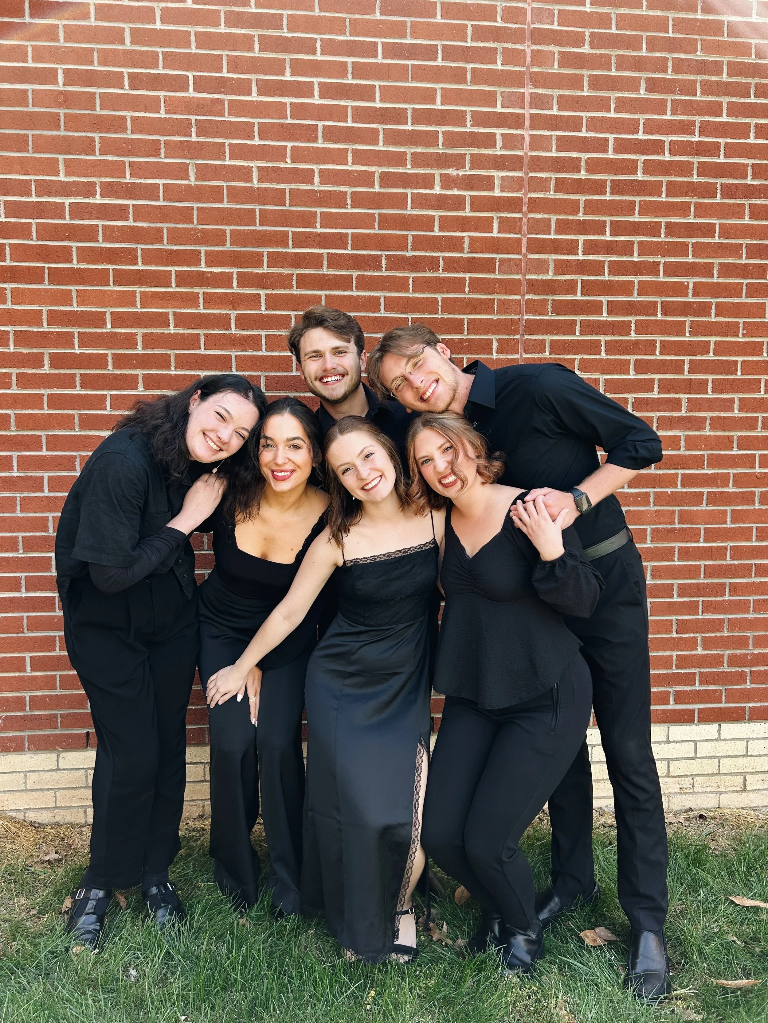 Group of six young adults dressed in black, standing outdoors in front of a brick wall, smiling and posing closely together.