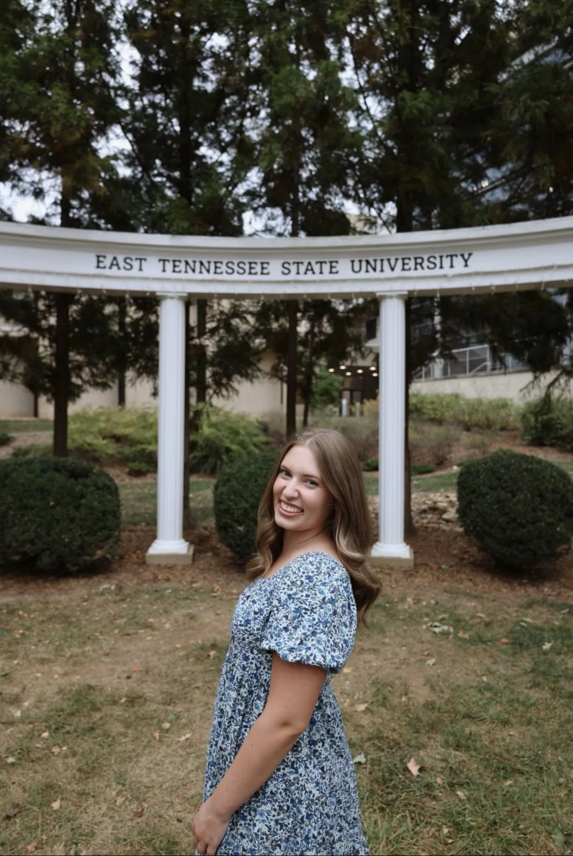 A young woman with long brown hair smiling in front of a sign that reads 'East Tennessee State University', surrounded by trees and bushes.