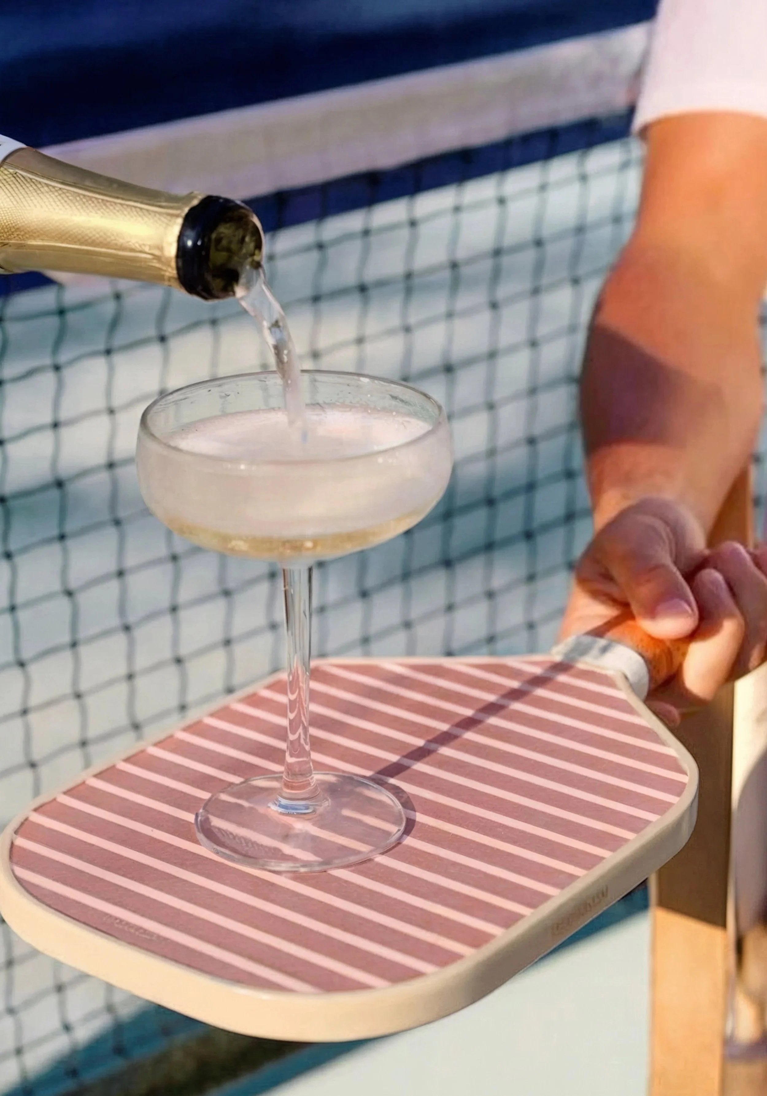 A person pouring champagne into a coupe glass on a serving tray held outdoors near a tennis court.