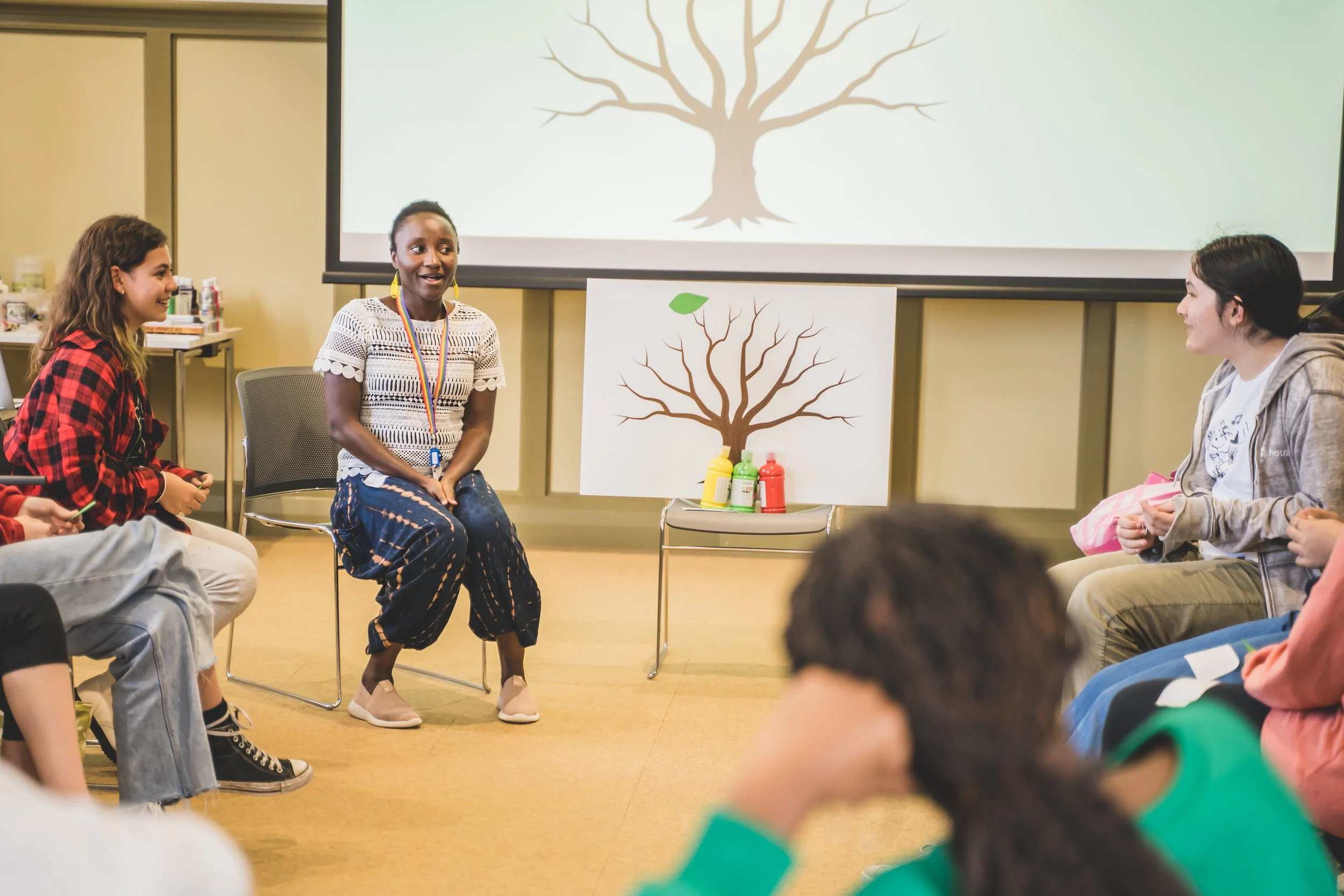 Wellbeing workshop featuring Hawa with a board, with a group of people surrounding her. The board has a tree with branches and no leaves on it
