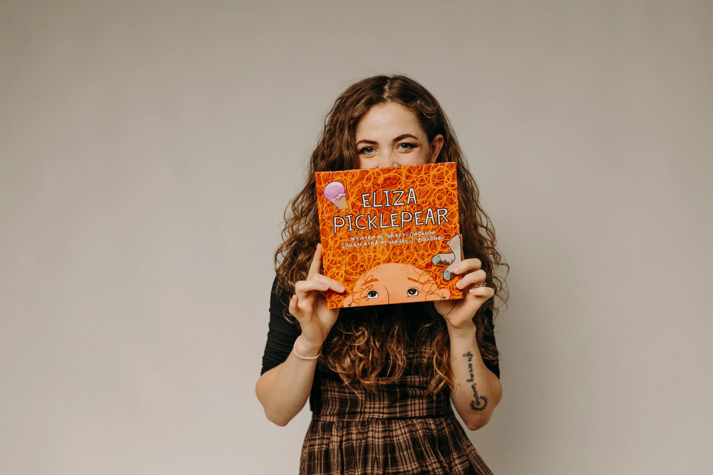 A woman with long curly hair holding a children's book titled "Eliza Picklepearl" in front of her face. The book cover features an illustration of a girl with curly hair and colorful design elements.