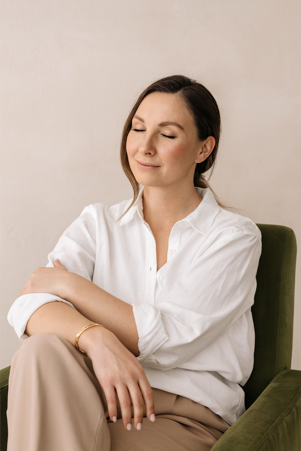 A woman with brown hair and a white blouse sitting with her eyes closed and a peaceful expression, arms crossed, on a green chair against a plain wall.