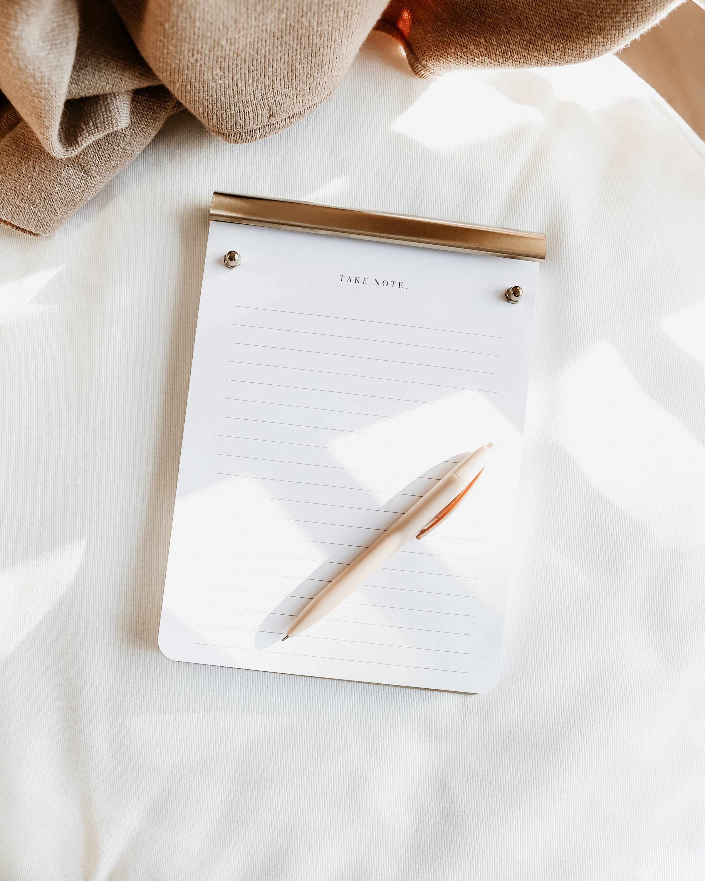 A lined notepad labeled 'Take Note' with a pen on a white surface, partially covered by a beige fabric.