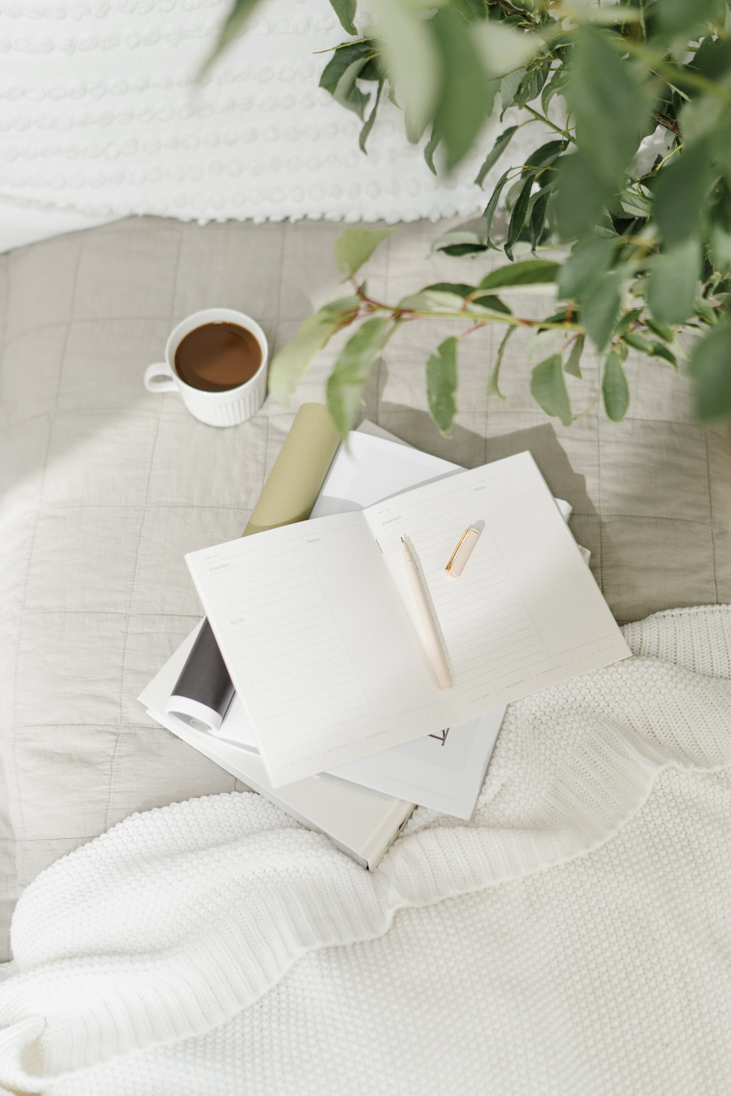 Open planner, pen, and glasses on a bed with a cup of coffee, a rolled-up poster, and a potted plant nearby.