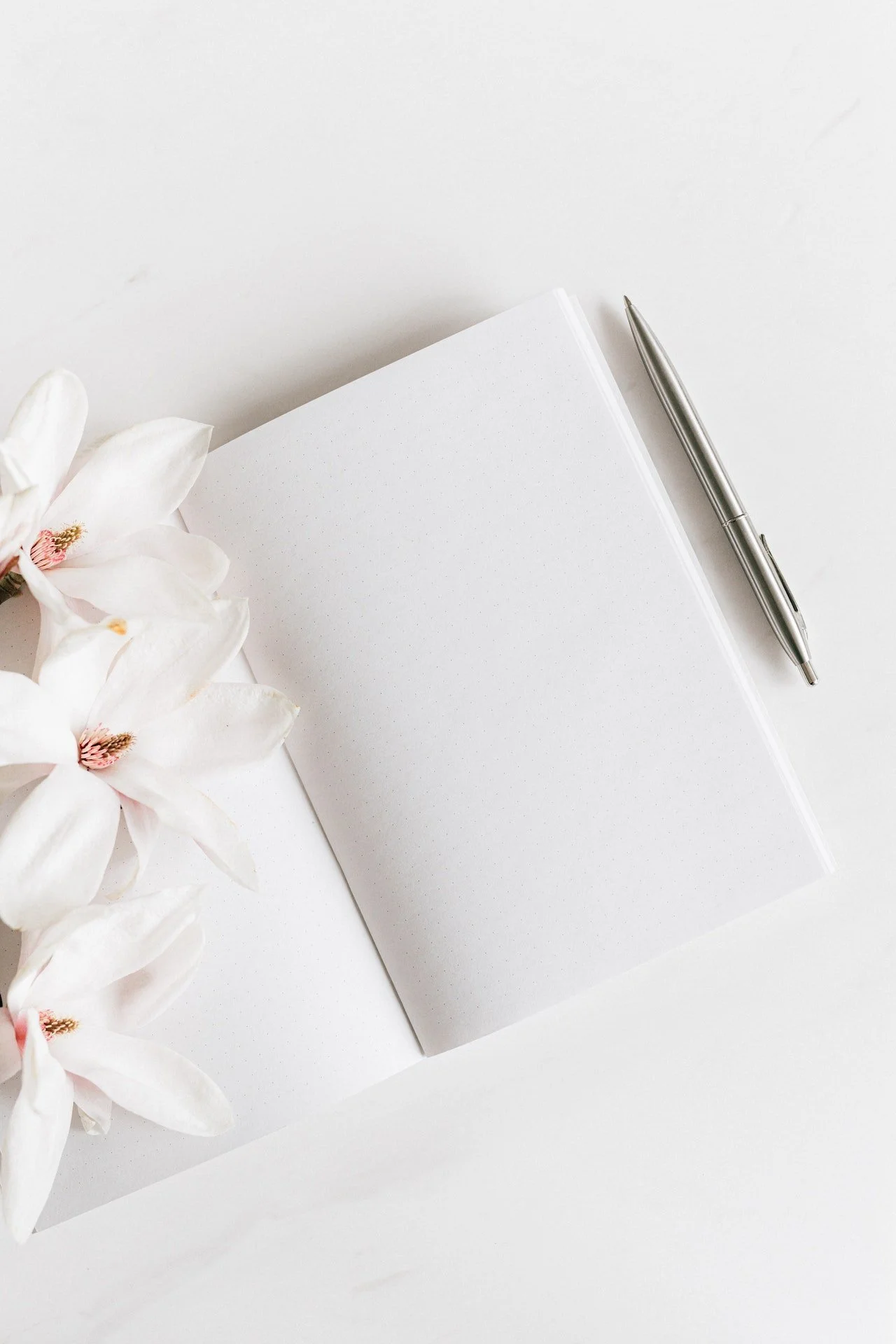 White notebook with dotted pages, a silver pen, and white flowers on a white background.