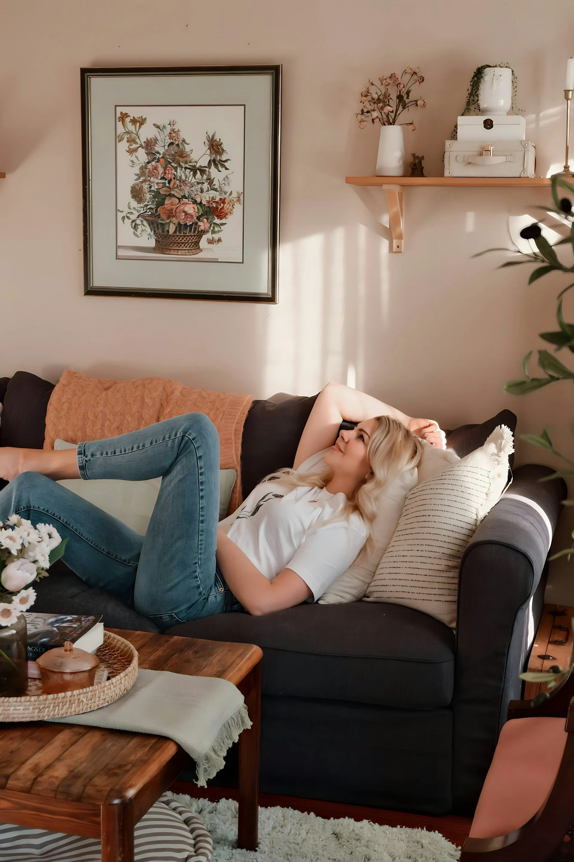 A young woman with blonde hair is lying on a dark-colored sofa, smiling and relaxed with her arms behind her head, in a cozy living room.