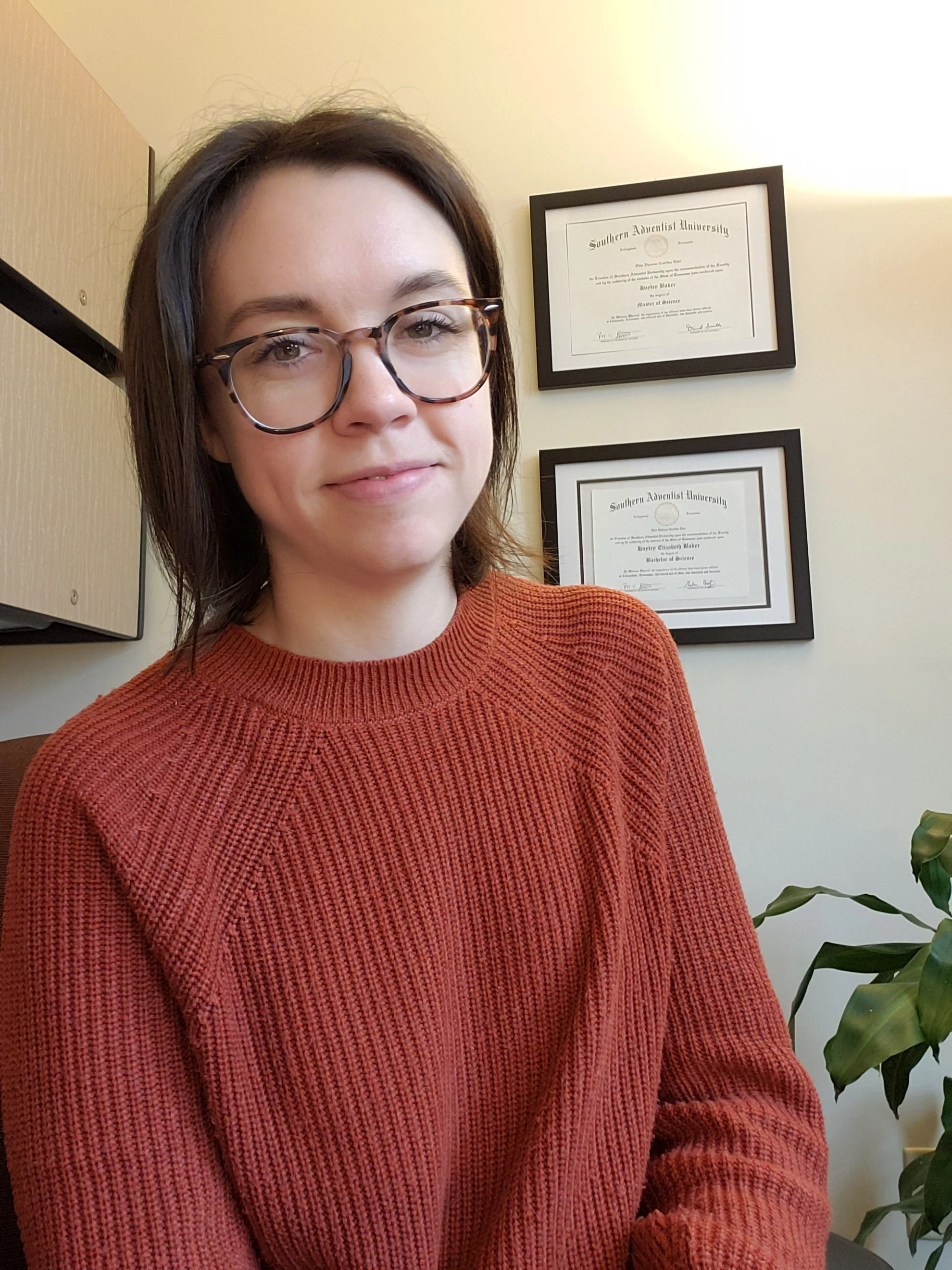 A woman with brown hair and glasses wearing an orange sweater, sitting indoors with framed diplomas or certificates hanging on the wall behind her.