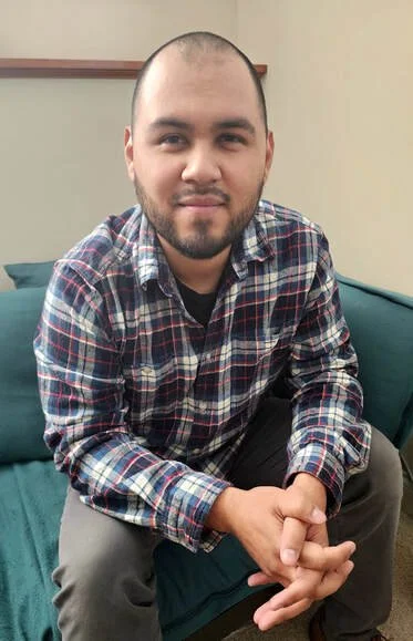 Young man with a beard and short hair sitting on a teal sofa, wearing a plaid shirt and gray pants, in an indoor setting.