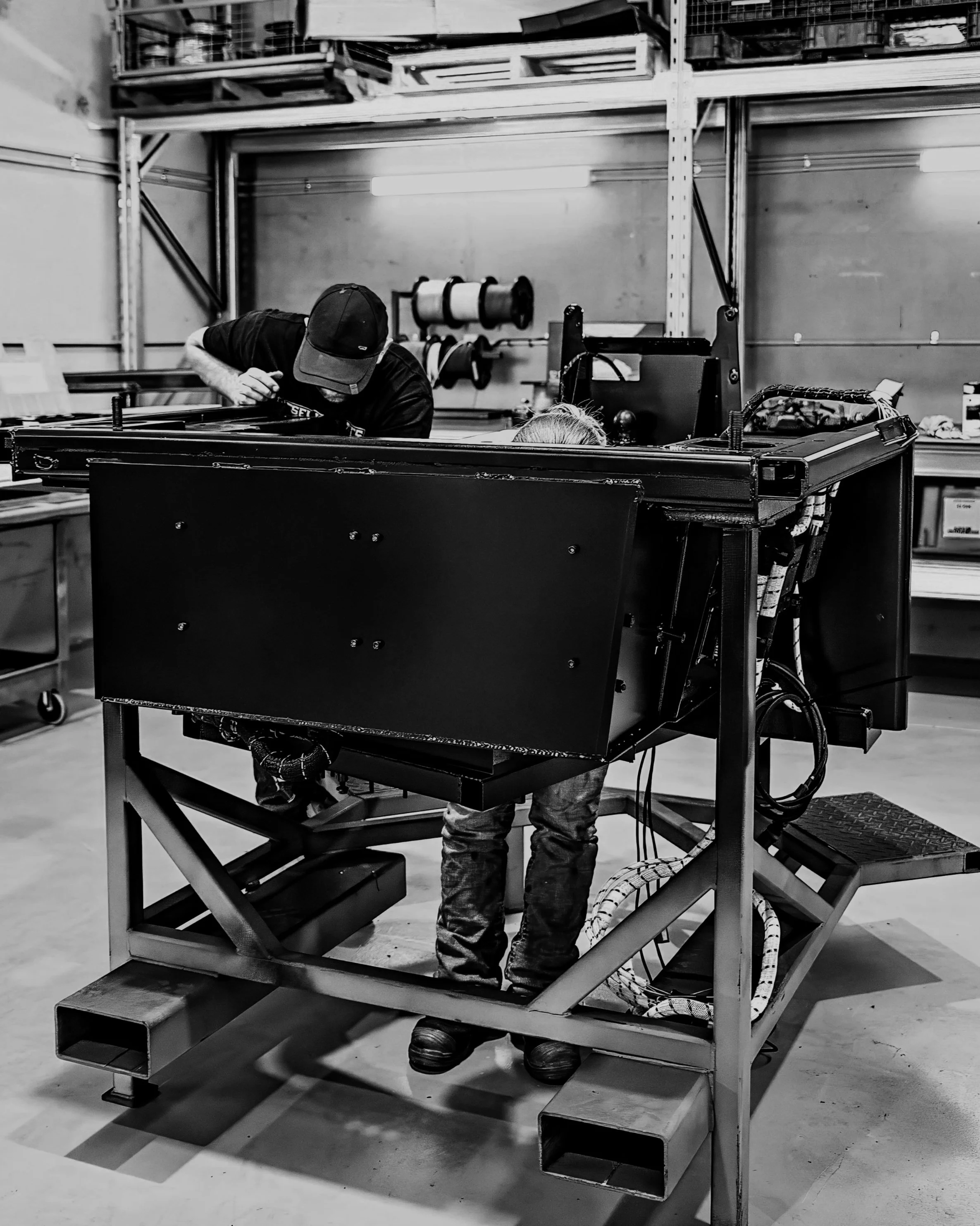 Two people working on a large metal machine or frame in an industrial workshop with shelves and spools of material in the background.