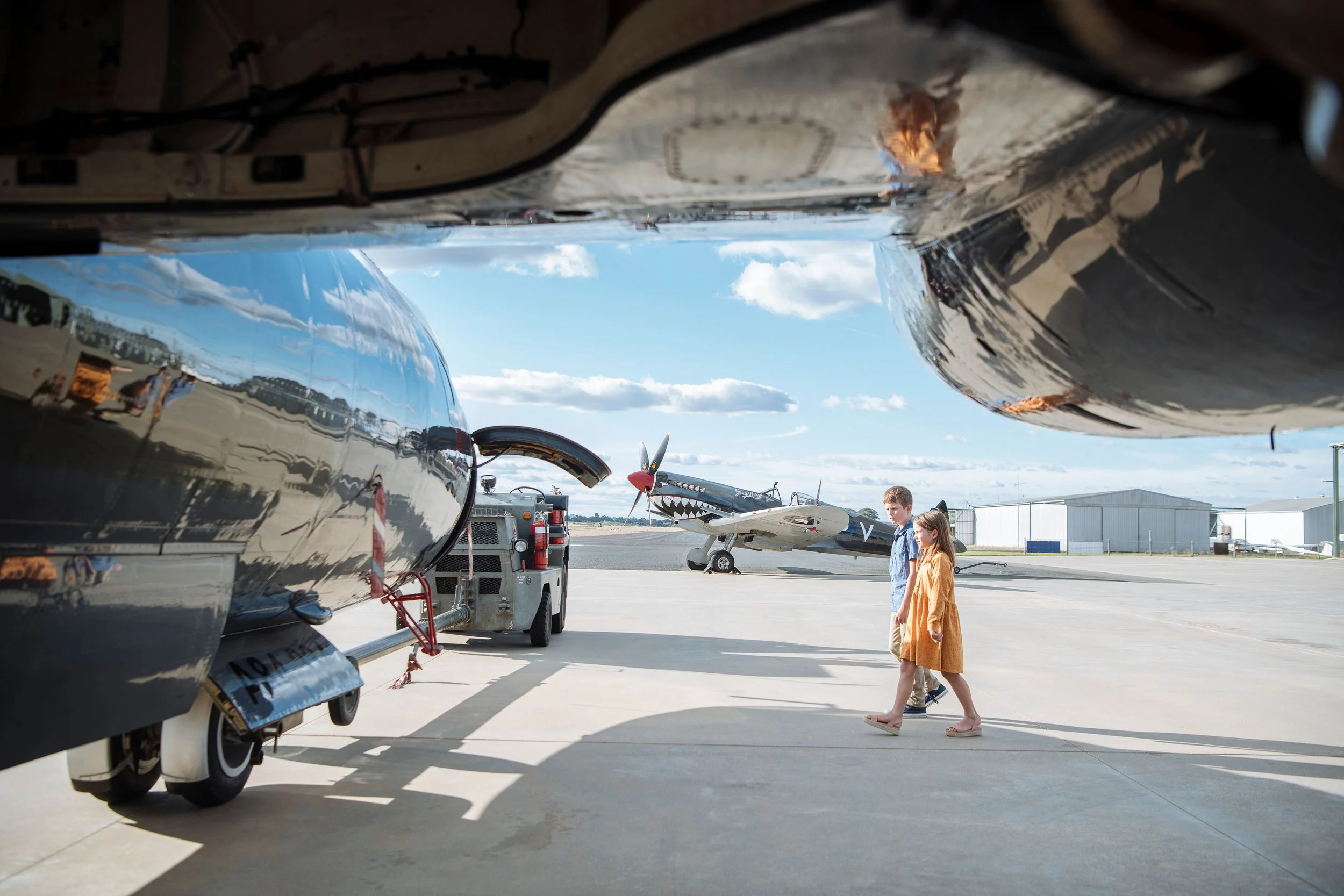 Two children walking at Temora Aviation Museum airfield with vintage airplanes, including one with shark mouth nose art, under a partly cloudy sky.