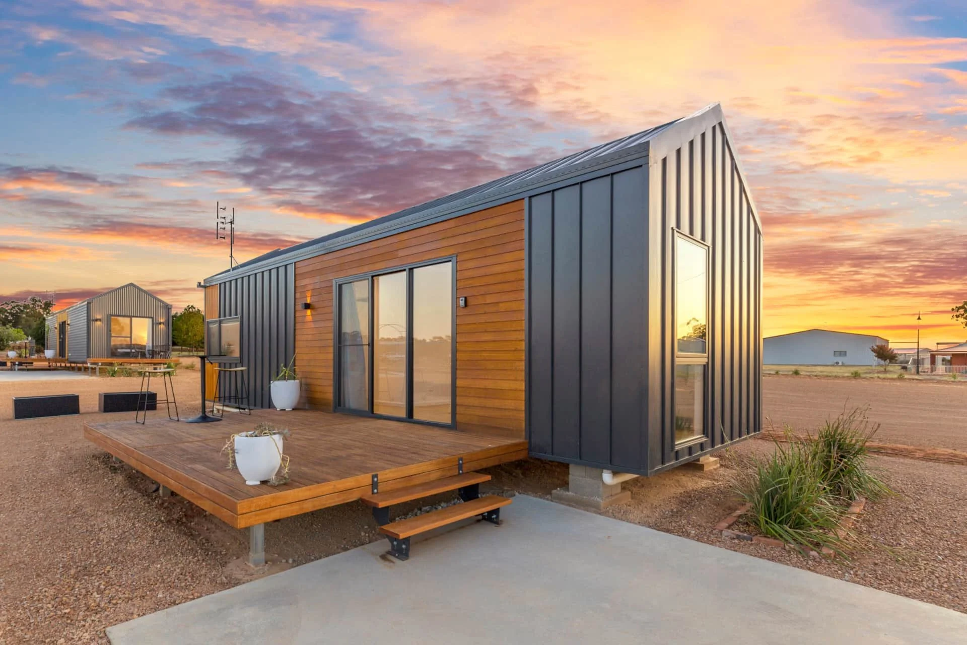 Modern tiny house on a gravel lot with a wooden deck, potted plants, and glass sliding doors, sunset sky in the background.