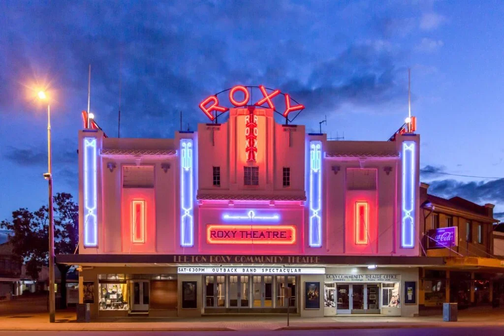 The Roxy Theatre at night with neon lights in pink and blue, a marquee with upcoming show details, and a theater sign on the top of the building.