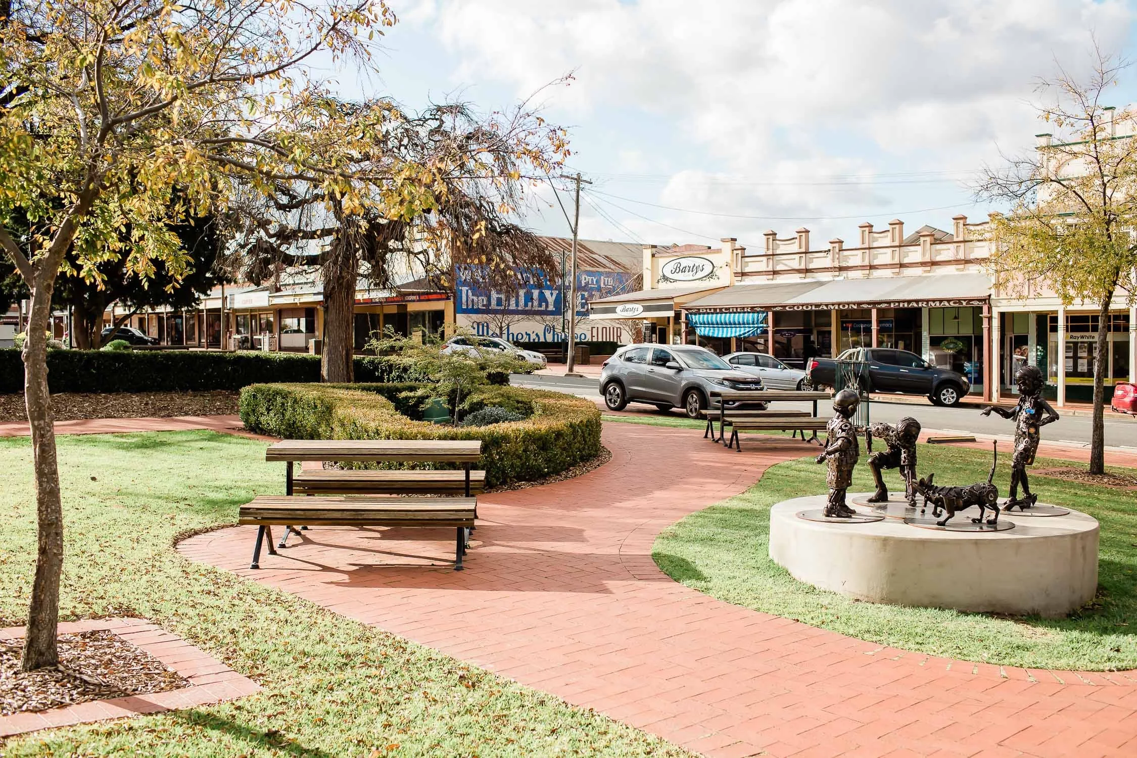 A small park with brick pathways, benches, trees, and a circular sculpture with children and dog statues at the center, in front of a row of storefronts on a street.