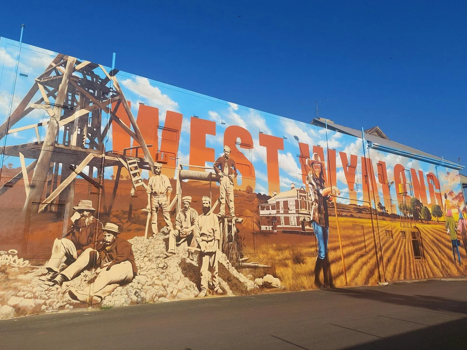 A mural in West Wyalong depicting historical figures, farming scenes, and the word 'WEST WYALONG' in large orange letters against a blue sky.