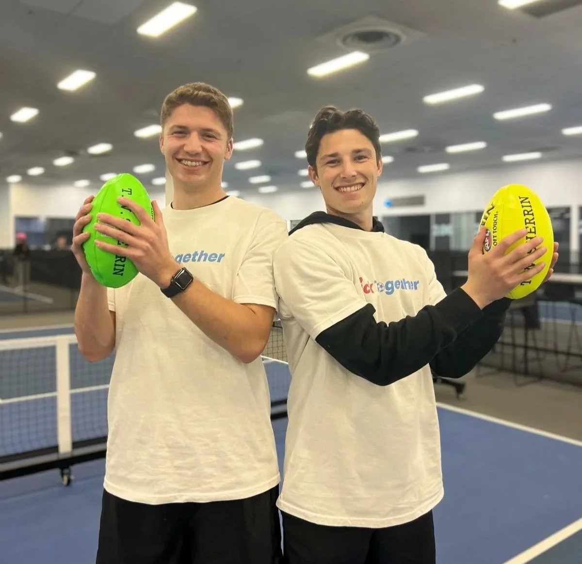 Coaches Dan and Ben holding green and yellow footballs at the indoor pickleball facility.