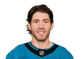 A young man with dark hair and a beard smiling, wearing a blue sports jersey against a white background.