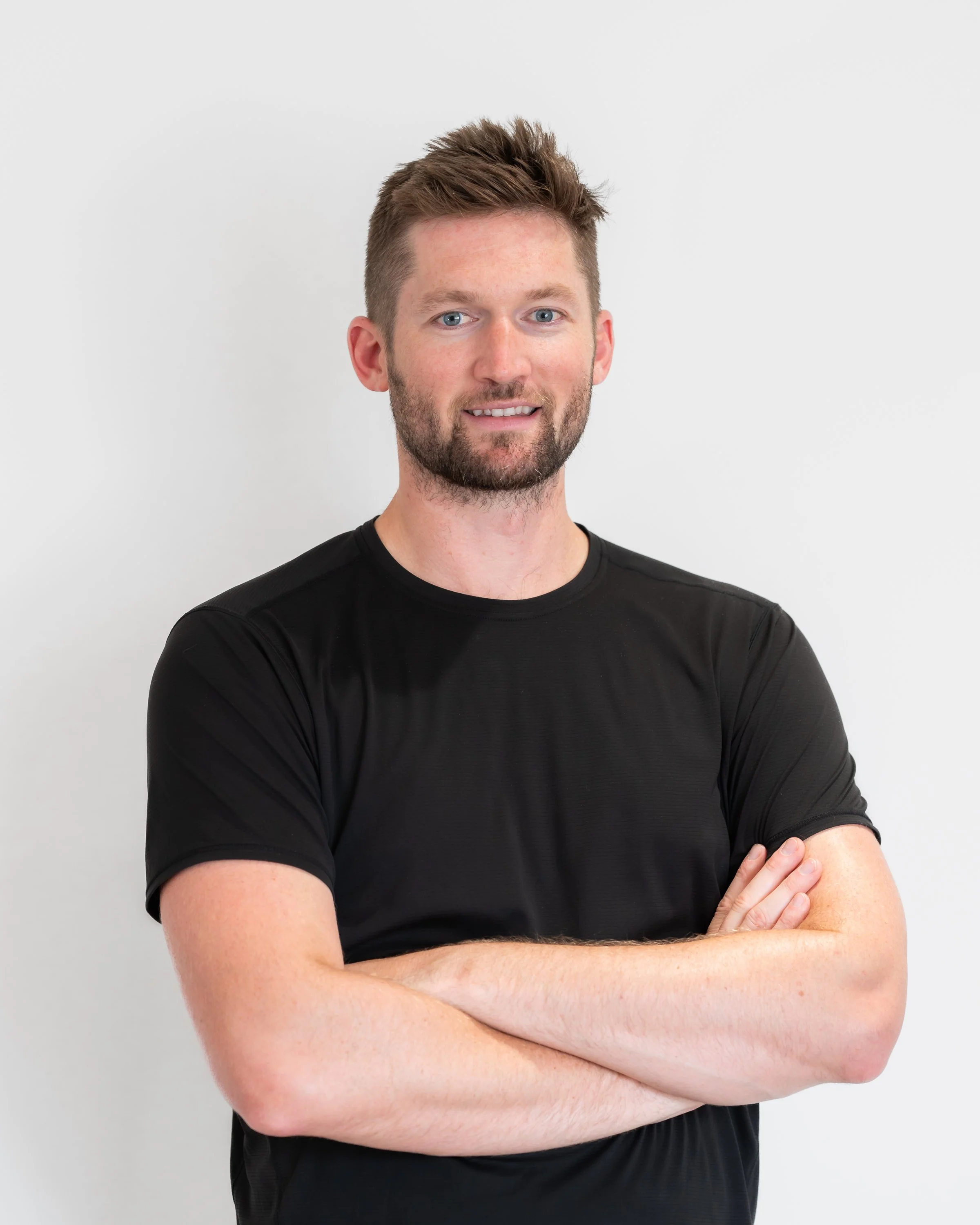 A man in black athletic wear sitting in a therapy or rehab room with a logo on the wall behind him that reads 'Sports Rehab'.