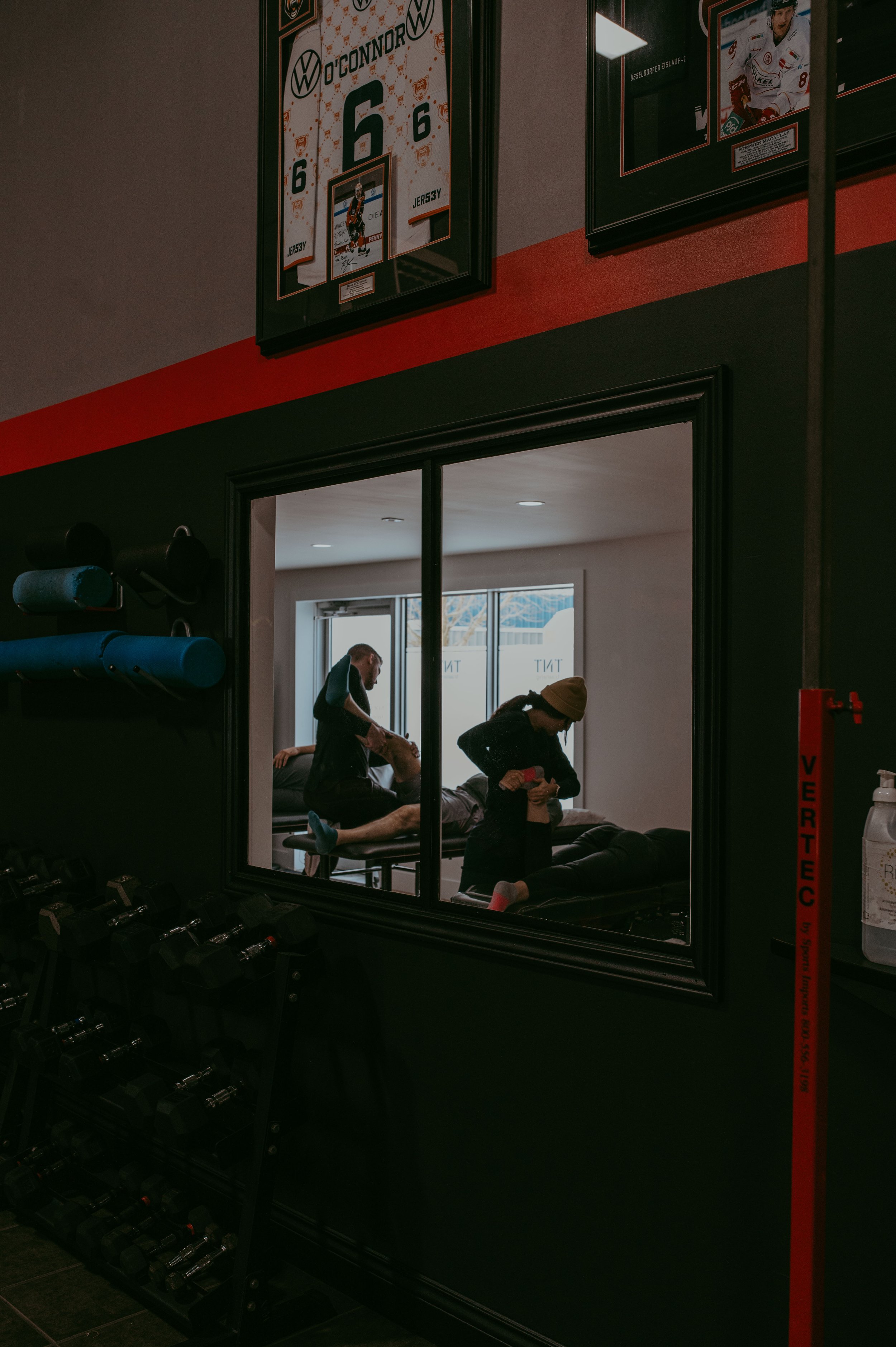 View through a window into a physiotherapy or massage room where a therapist is working on a patient lying face down on a treatment table, with another person beside them. The room is well-lit with window lighting and has fitness equipment and framed sports memorabilia on the walls.