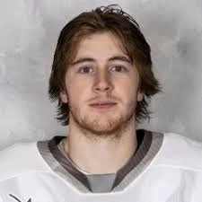 Portrait of a young man with shoulder-length brown hair, wearing a white sports jersey with gray trim, standing against a gray background.