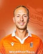 A man wearing an orange sports jersey with a badge, smiling, against an orange background.