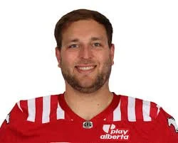 A man with brown hair, a beard, and a mustache, smiling, wearing a red football jersey with white stripes on the sleeves and logos.