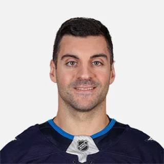 A man with short dark hair and a light beard, smiling, wearing a dark sports jersey with blue and silver accents and an NHL logo on a white background.