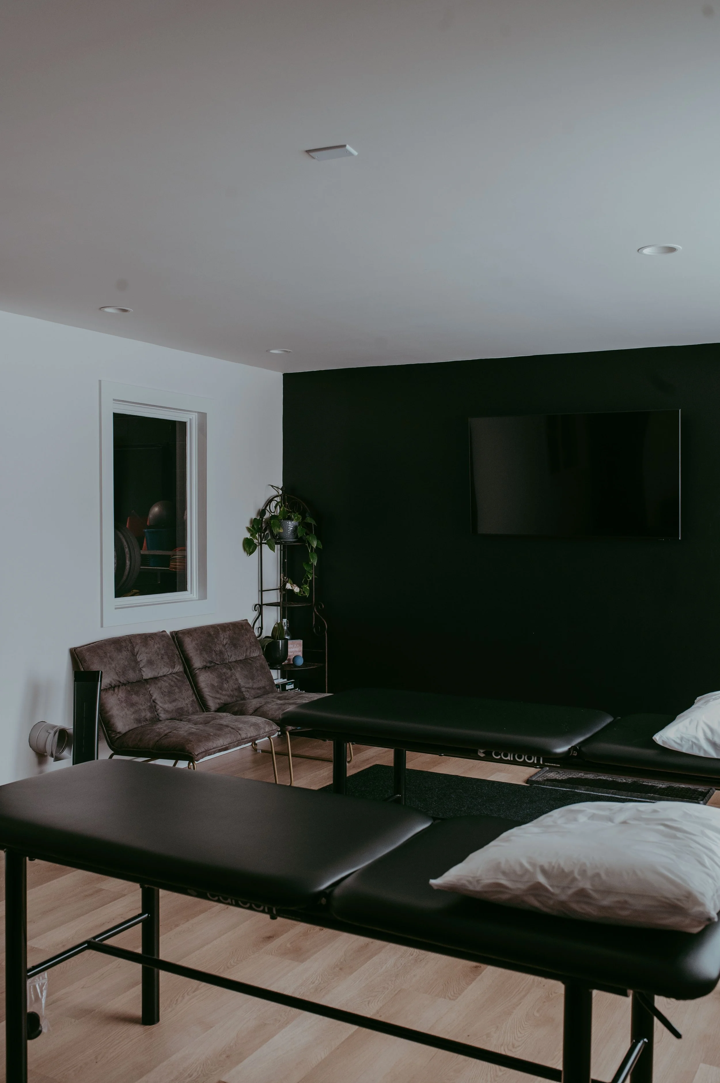 A room with two treatment tables, a black wall-mounted TV, a brown chair, a window, and a small black shelf with a plant.