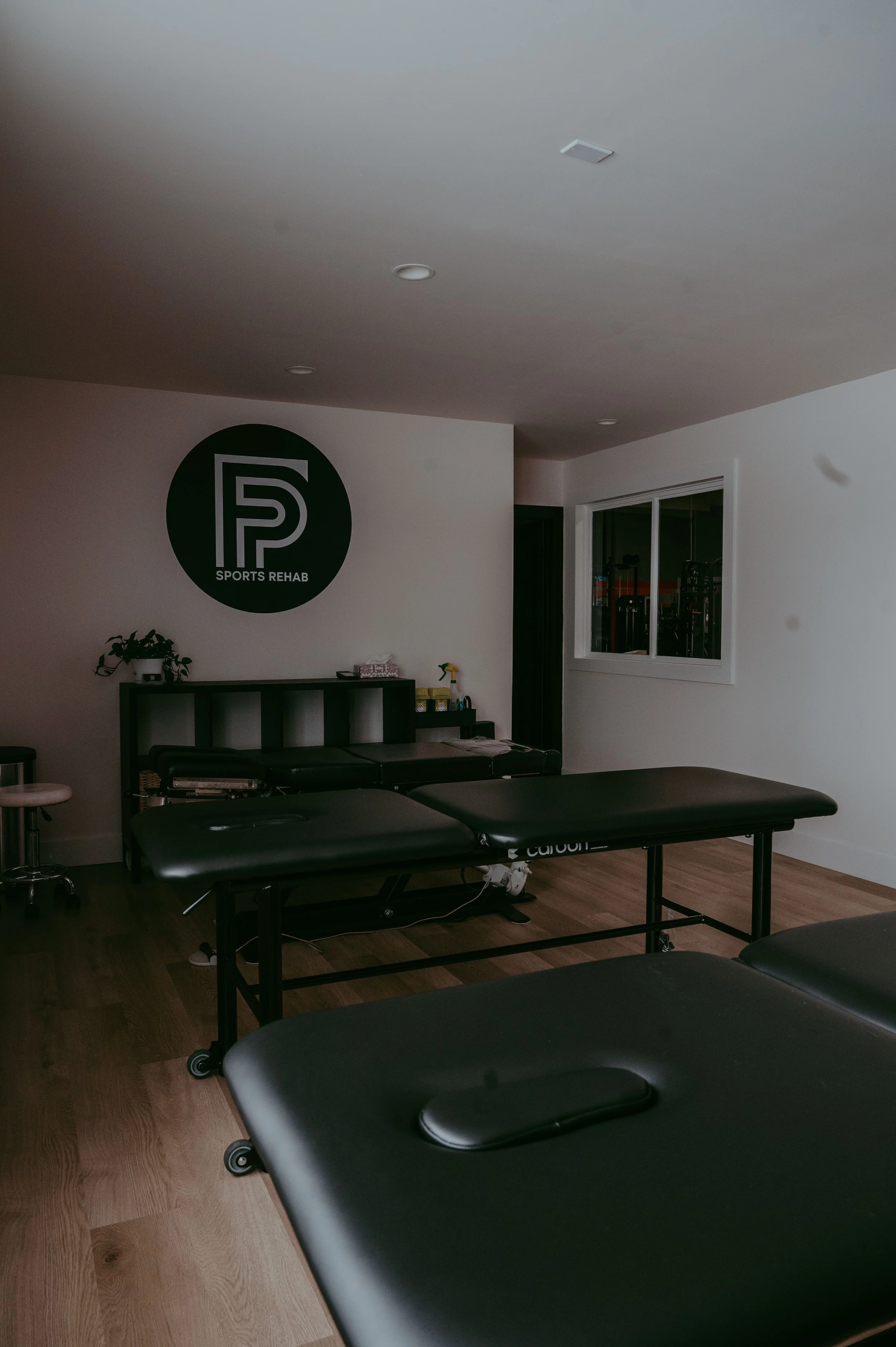 Empty physical therapy or sports rehabilitation treatment room with treatment tables, a black logo on the wall reading 'SPORTS REHAB,' and a window showing gym equipment outside.