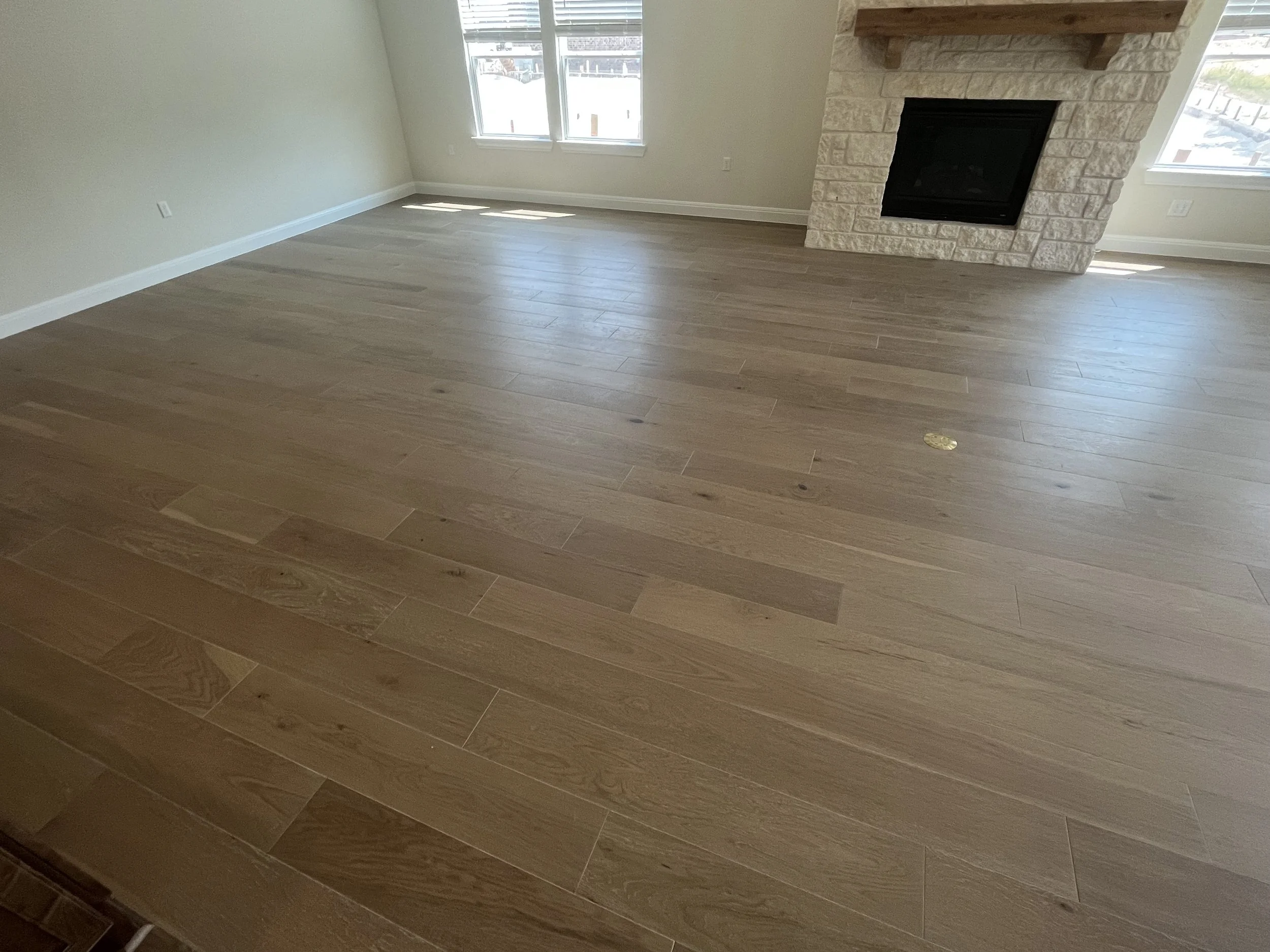 Empty living room with natural light, beige walls, hardwood floors, dual large windows, and a stone fireplace with a wooden mantel.