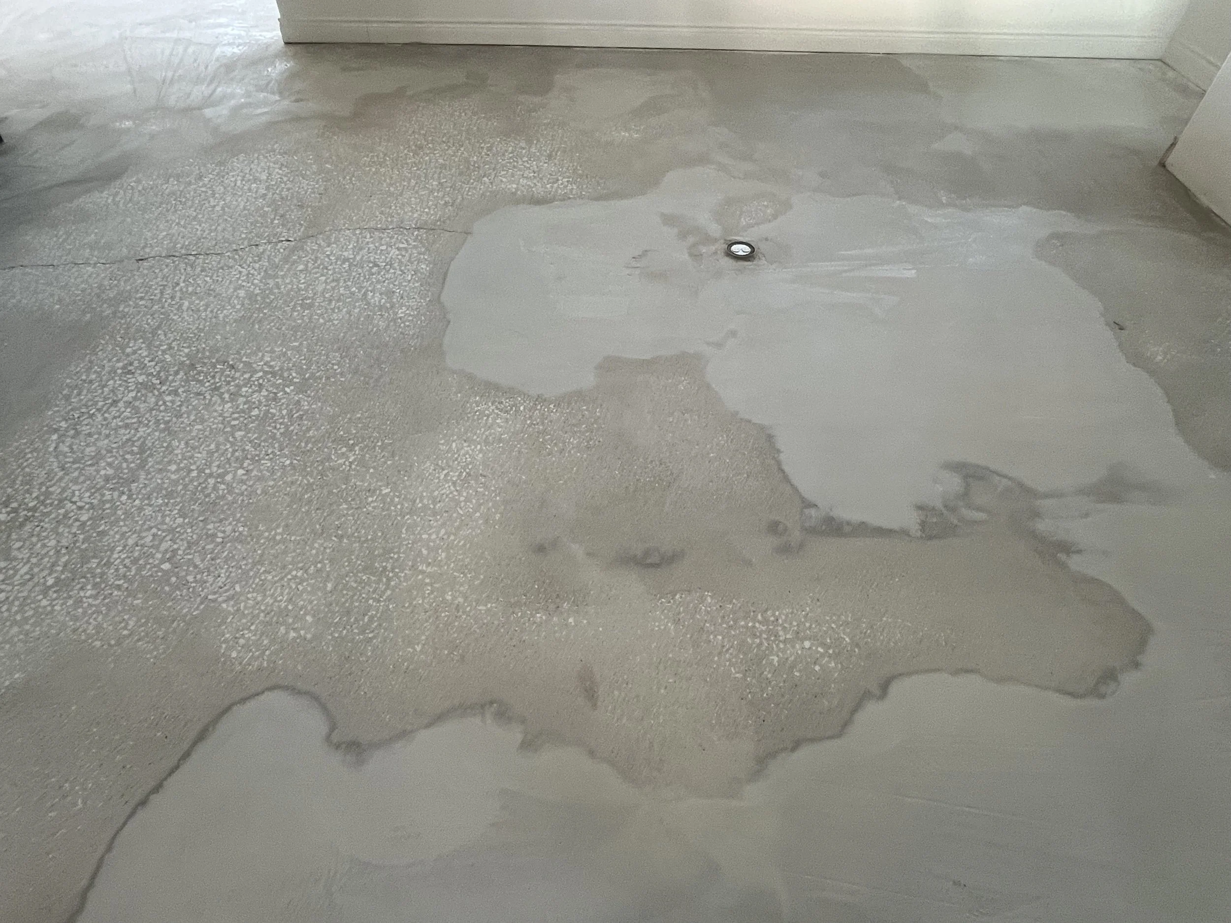 Unfinished concrete floor with patches of gray and white, and a floor drain in the center, in a room with white walls.