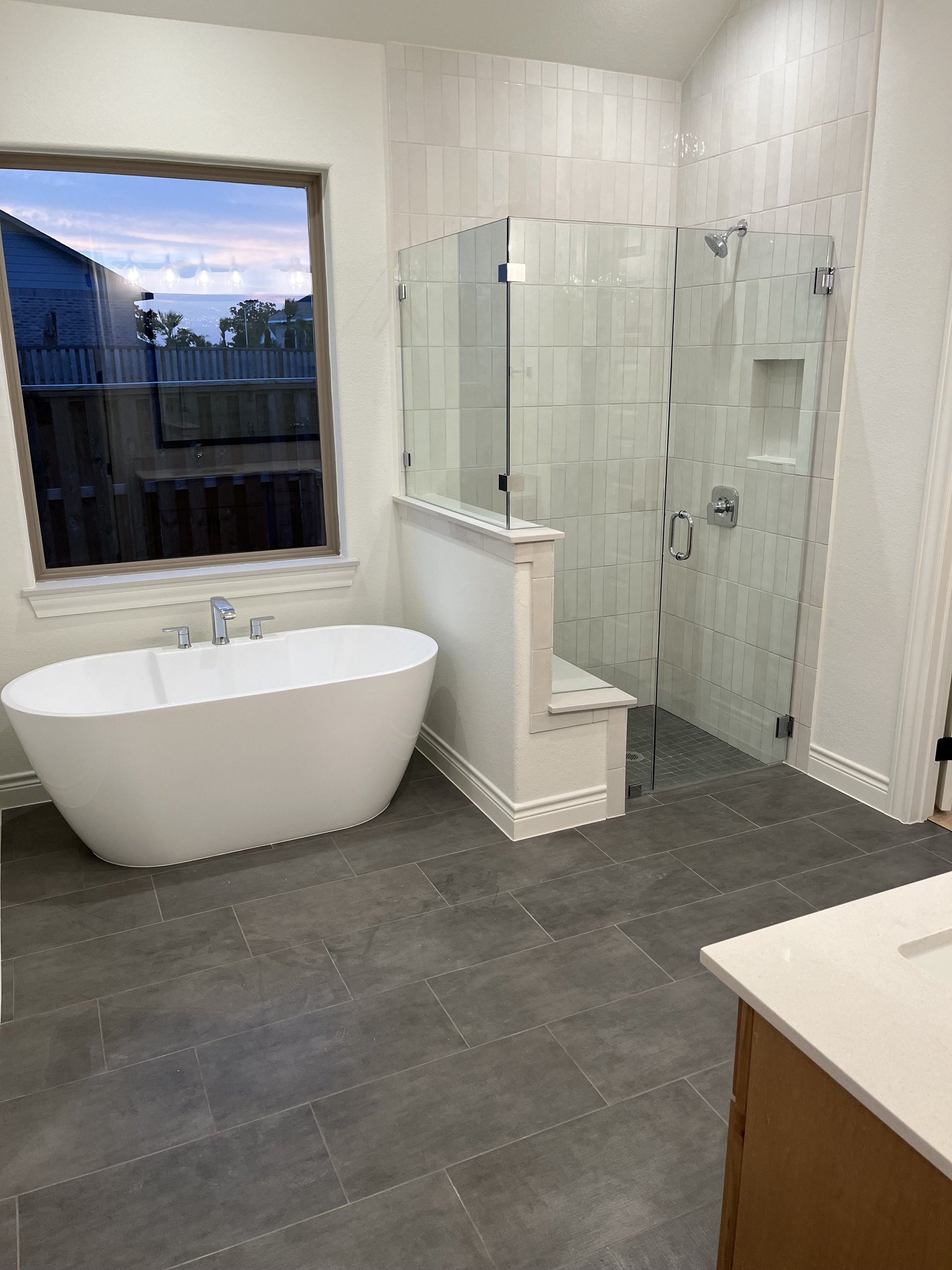 Modern bathroom with a white freestanding bathtub, a glass-enclosed shower with tiled walls, a window showing an outdoor view, and a sink with a wooden cabinet.