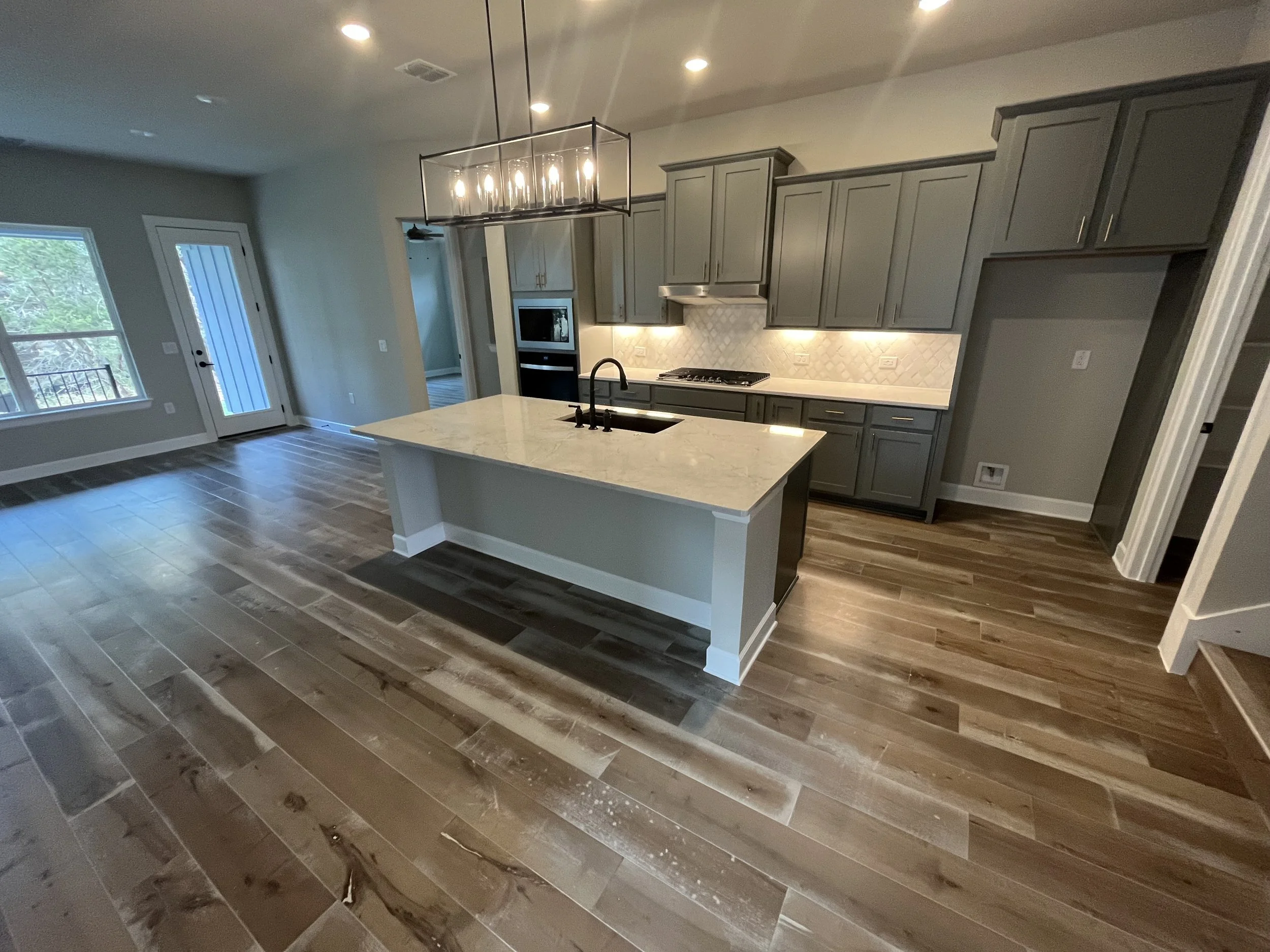 Open-concept kitchen with gray cabinets, white marble island, wood flooring, and modern lighting, overlooking a living area with large windows and a door, leading to an outdoor space.