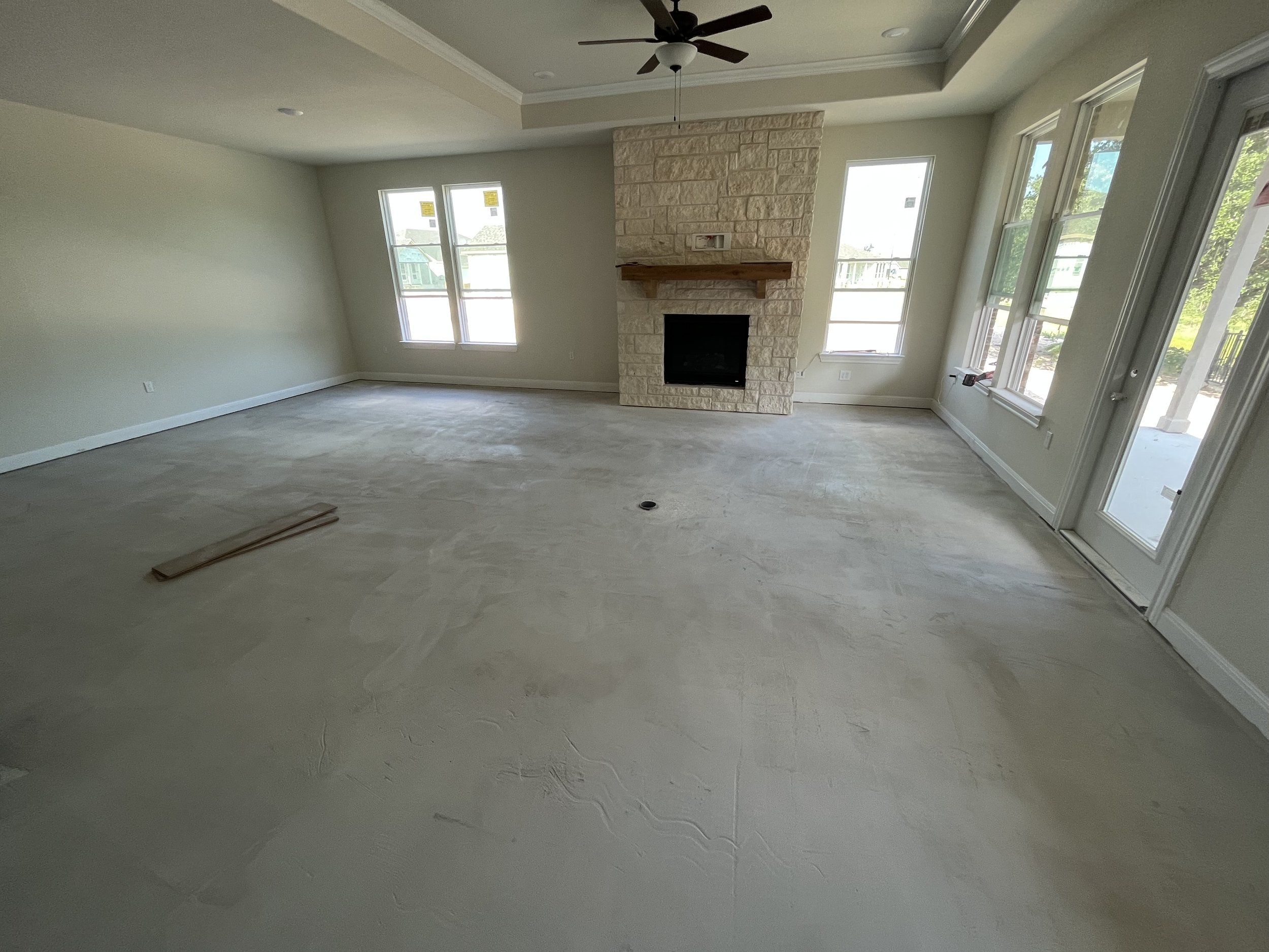 Empty living room with large windows, a stone fireplace with a wooden mantel, ceiling fan, and a concrete floor.