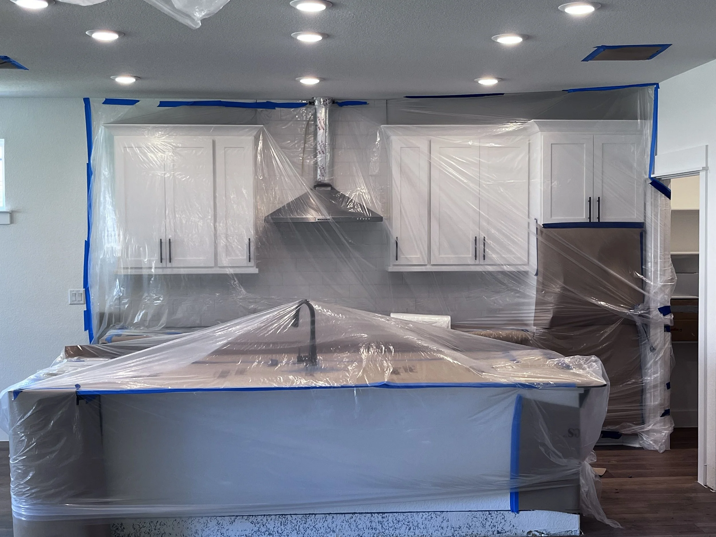 Kitchen under renovation covered with protective plastic sheeting and blue painter's tape, with white cabinets, a range hood, and a kitchen island.