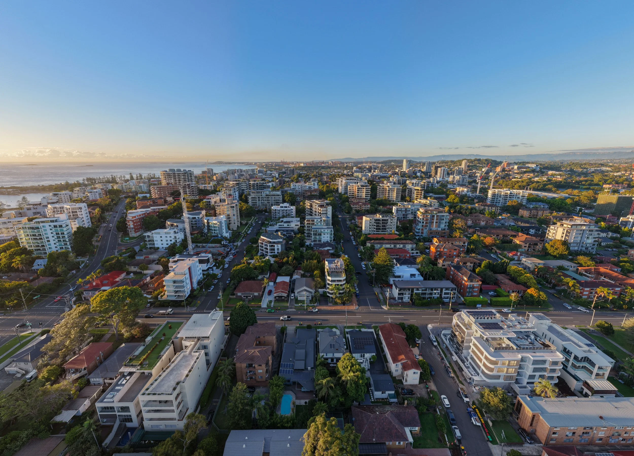 Aerial view of a city with a mix of modern and traditional buildings, streets, trees, and a body of water in the background, under a clear blue sky.