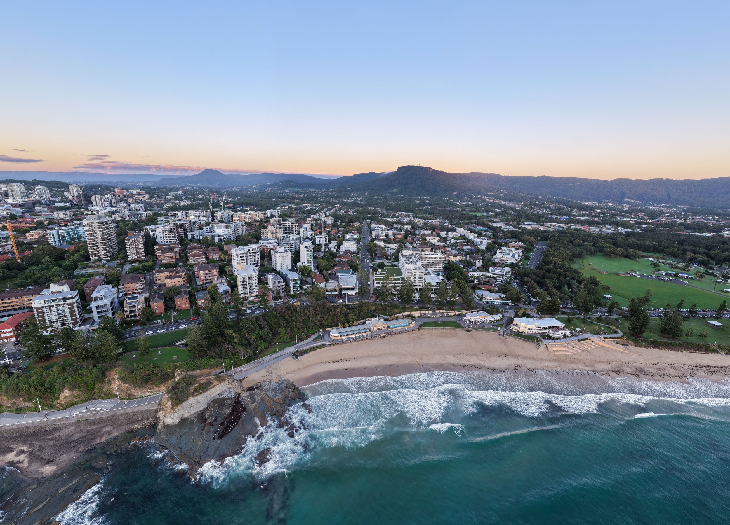 Aerial view of a city along the coast at sunset, with buildings, green parks, and a sandy beach with waves crashing, and mountains in the background.