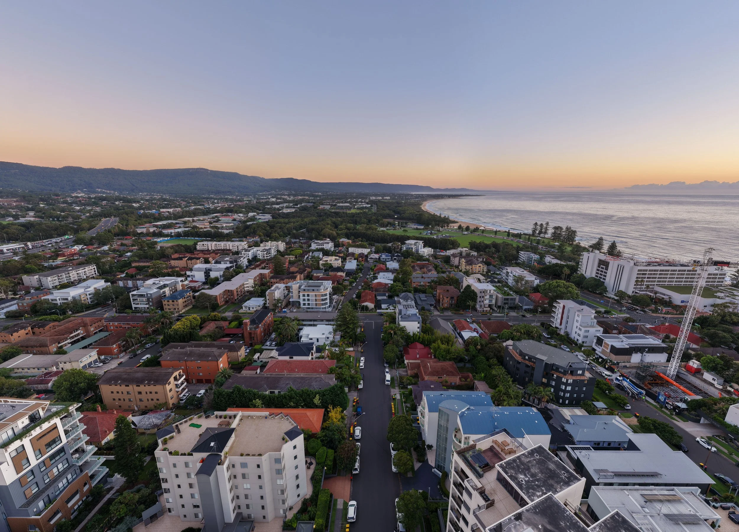 Aerial view of a coastal city at sunset, showing residential and commercial buildings, streets, greenery, and the ocean in the background.