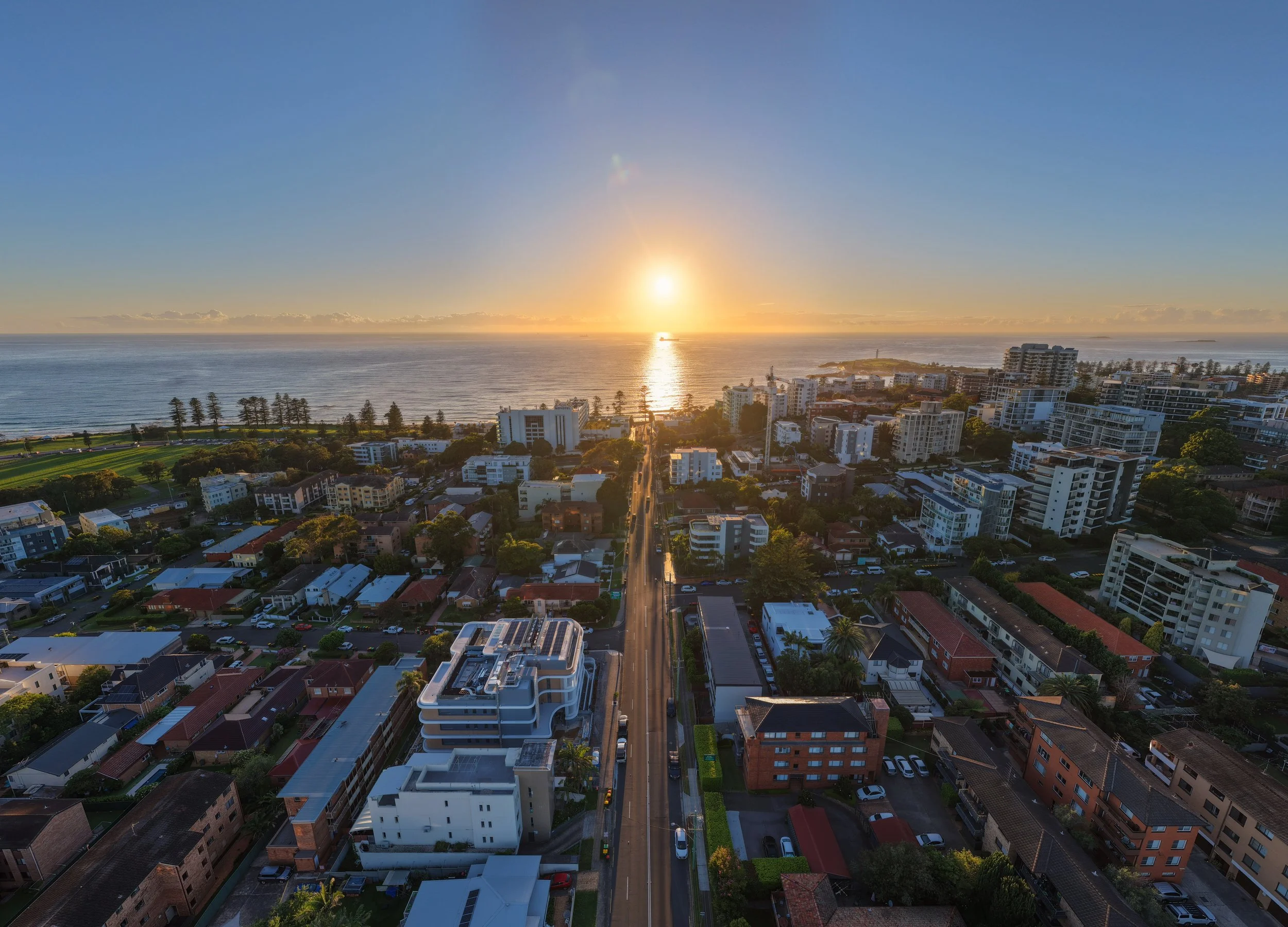 Aerial view of a city at sunset with a clear sky, overlooking the ocean, with buildings, trees, and streets.