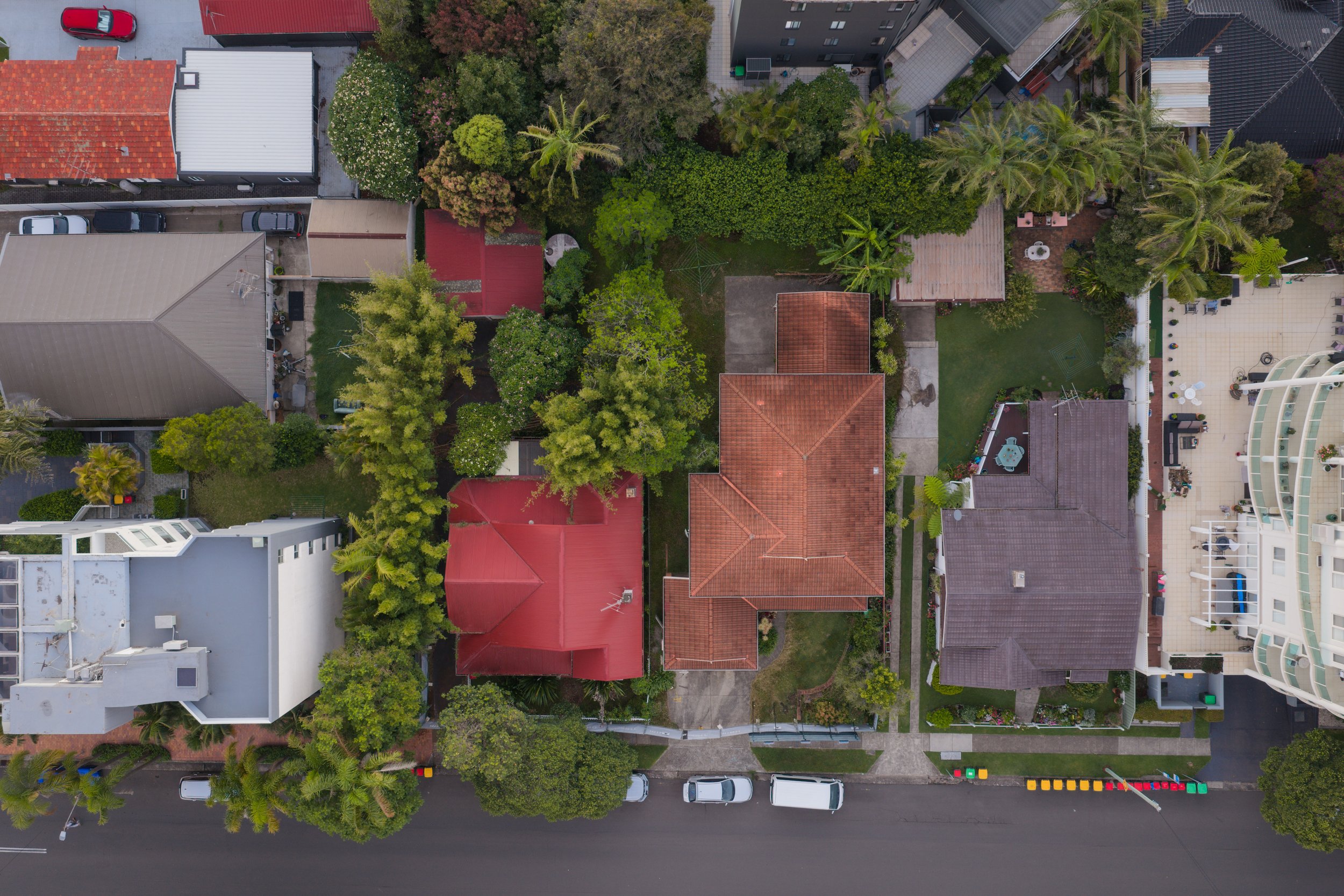 Aerial view of residential houses with red roofs, green trees, and a street with parked cars.