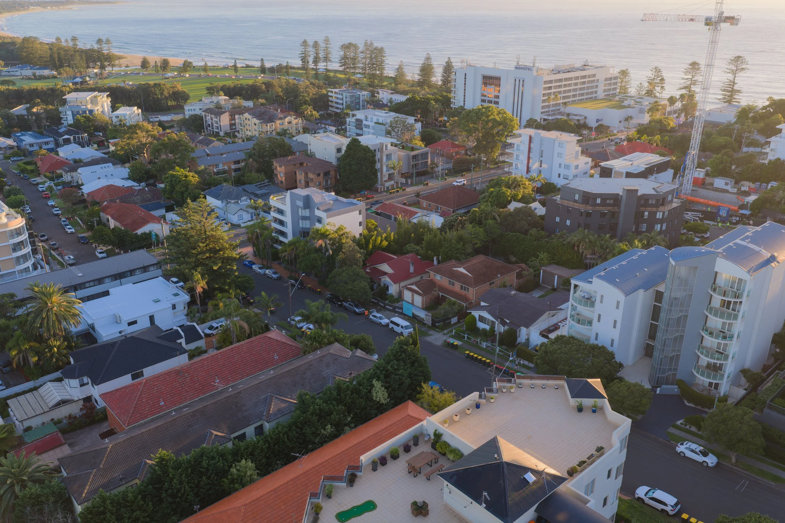 Aerial view of a coastal cityscape at sunset, showing a mix of residential and hotel buildings surrounded by greenery, with the ocean in the background.