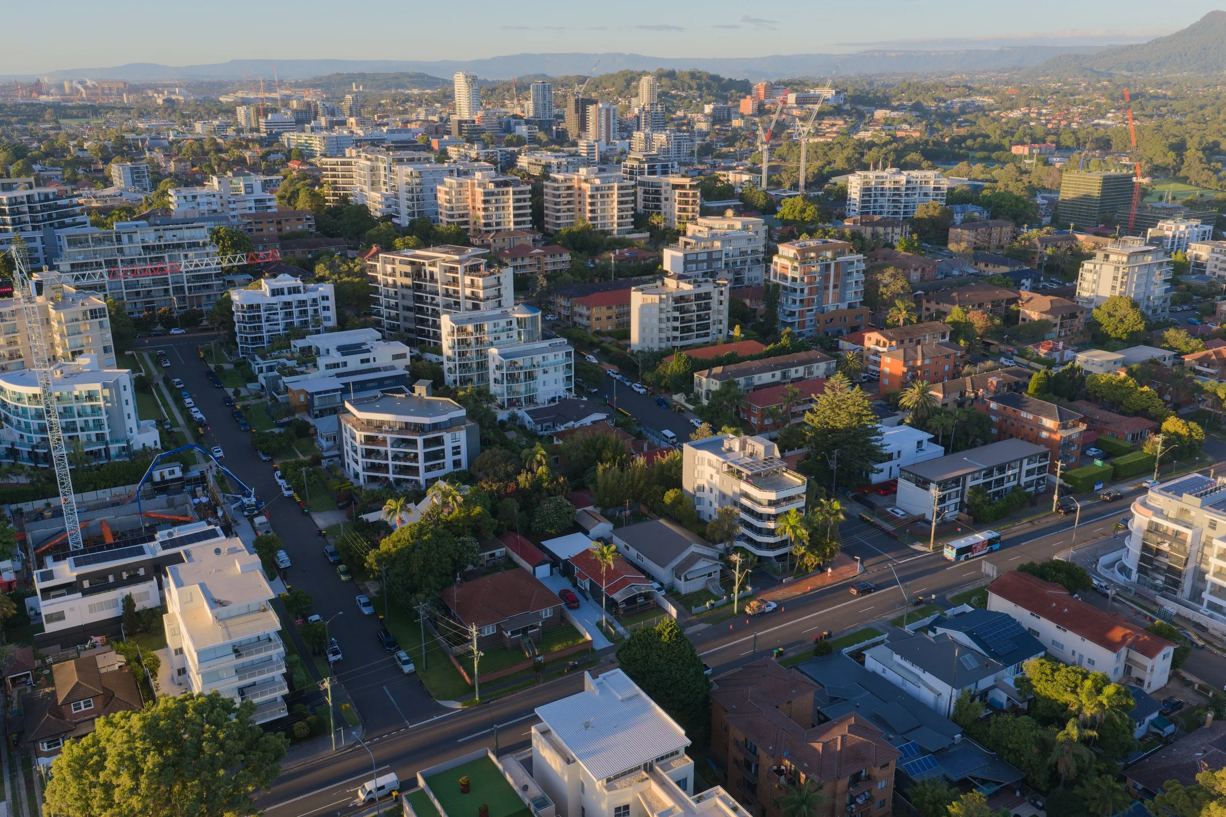 Aerial view of a cityscape with modern apartment buildings, residential houses, and streets, with low hills in the background during sunset.